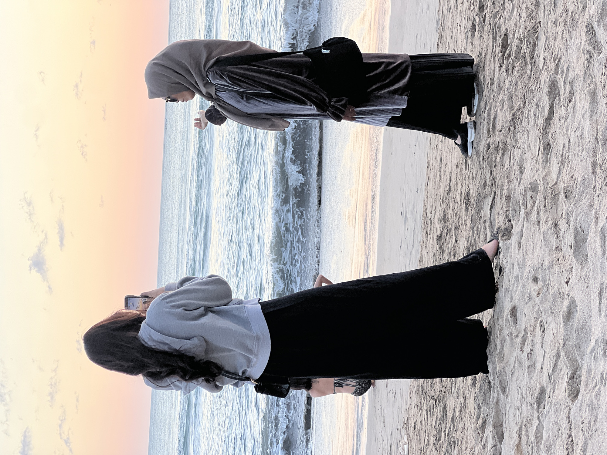 Two women are standing on a sandy beach in Kuta, Indonesia, near the ocean at sunset. The woman on the left has long, dark hair and is wearing a gray hooded sweatshirt and a long black skirt. She is holding up a smartphone to take a picture of the ocean. She has a black purse slung over her shoulder. The woman on the right is wearing a hijab and sunglasses, and is wearing a long, dark gray velvet dress over a black skirt. She is holding her right hand up as if to feel the air or catch something. She has a black purse with a silver zipper on her right side. The ocean has small waves, and the sky is a soft pink and orange hue, indicating sunset. In the background, another person wearing a patterned bikini is partially visible, sitting on the sand. The sand is light brown and textured.