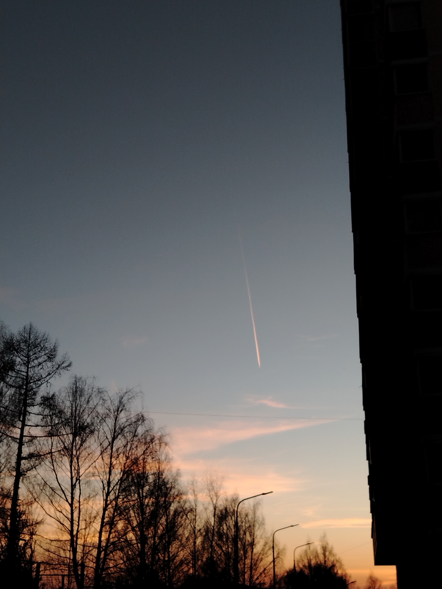 The image captures an evening sky in Izhevsk, Russia. The sky transitions from a deep gray-blue above to a warm orange-pink near the horizon, indicating sunset. A prominent contrail stretches across the sky. Silhouetted trees are at the bottom left and a dark, silhouetted building occupies the right side of the frame. Two streetlights are partly visible among the trees. There are no visible people.