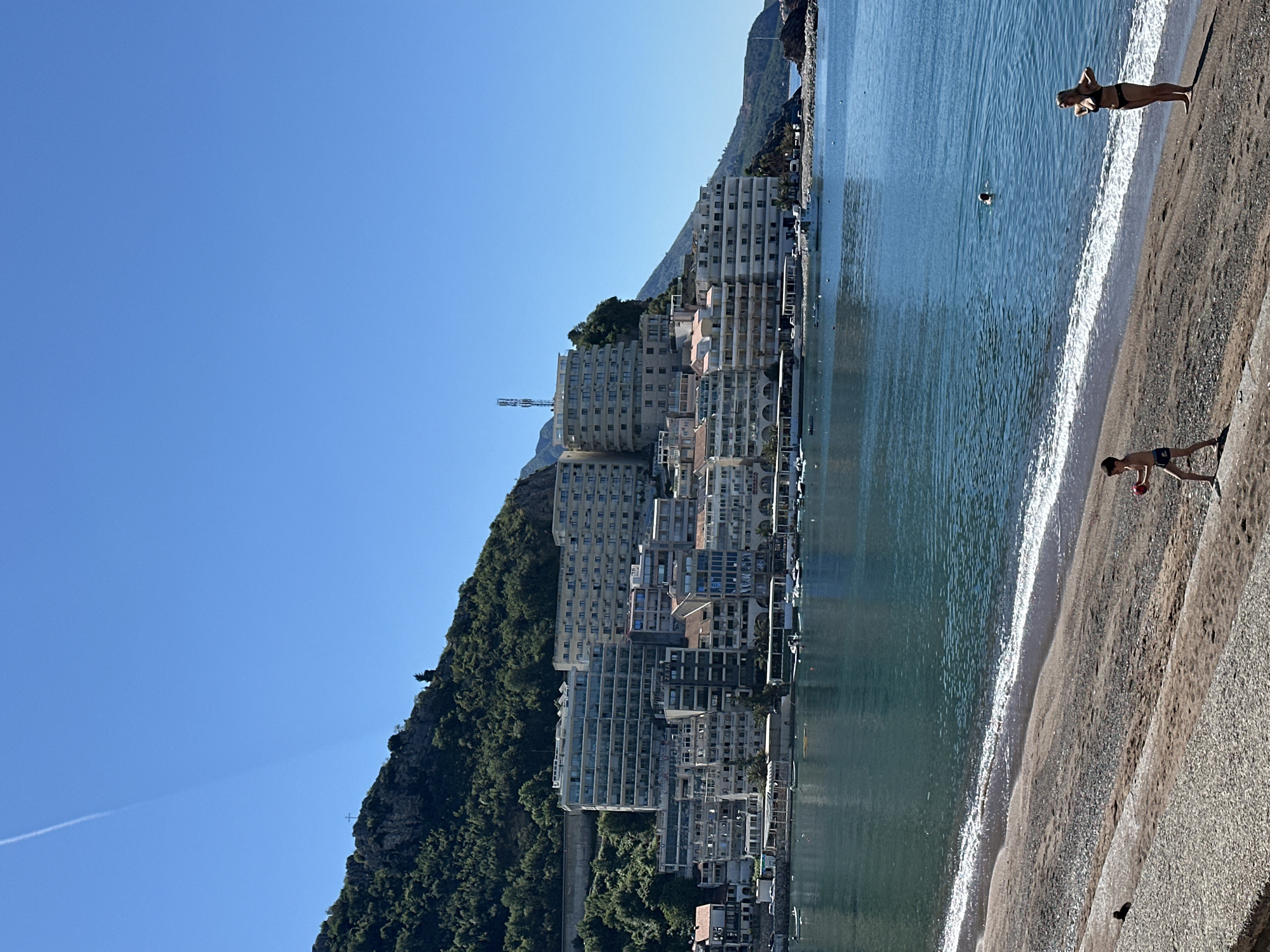The image shows a coastal scene in Boreti, Montenegro. A hillside covered with dense green vegetation slopes down to a line of high-rise buildings. The Adriatic Sea stretches alongside the buildings, with gentle waves breaking on a sandy beach. Two people are visible on the beach: one standing at the water's edge and another walking on the sand. A swimmer is visible in the water. The sky is a clear, cloudless blue.