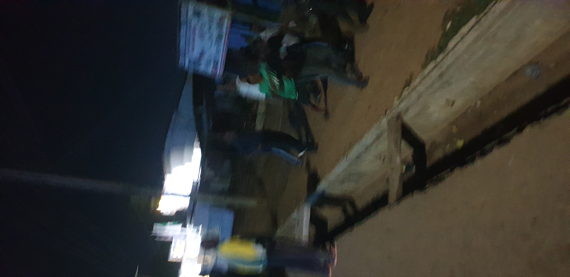 The image captures a dimly lit, blurry nighttime street scene, likely in an urban or semi-urban area such as Sabon Tasha, Nigeria. The perspective is somewhat elevated, looking down onto a street or pathway.
In the foreground and extending towards the center is an unpaved or dirt ground, suggesting a less developed road or a busy market area. Along the right side of the frame, there appears to be a structure, possibly a row of informal market stalls or a building, constructed with what looks like wooden planks or similar materials. This structure casts shadows onto the ground.
The scene is populated by several individuals, though most are indistinct due to the low light and motion blur. One person near the center is somewhat discernible, wearing a bright green top or garment. Other figures are present, but their details and activities are unclear beyond general movement or standing.
Bright artificial light sources are visible on the left side of the frame, illuminating parts of the scene and creating reflections. These lights suggest nearby shops, streetlights, or other establishments operating after dark. The overall lighting indicates that the photograph was taken at night.
The image quality is low, characterized by significant blur and graininess, which obscures fine details and makes it difficult to discern specific objects, facial expressions, or distinct actions. No legible text is visible anywhere in the frame. The atmosphere is one of a busy, nocturnal public space.