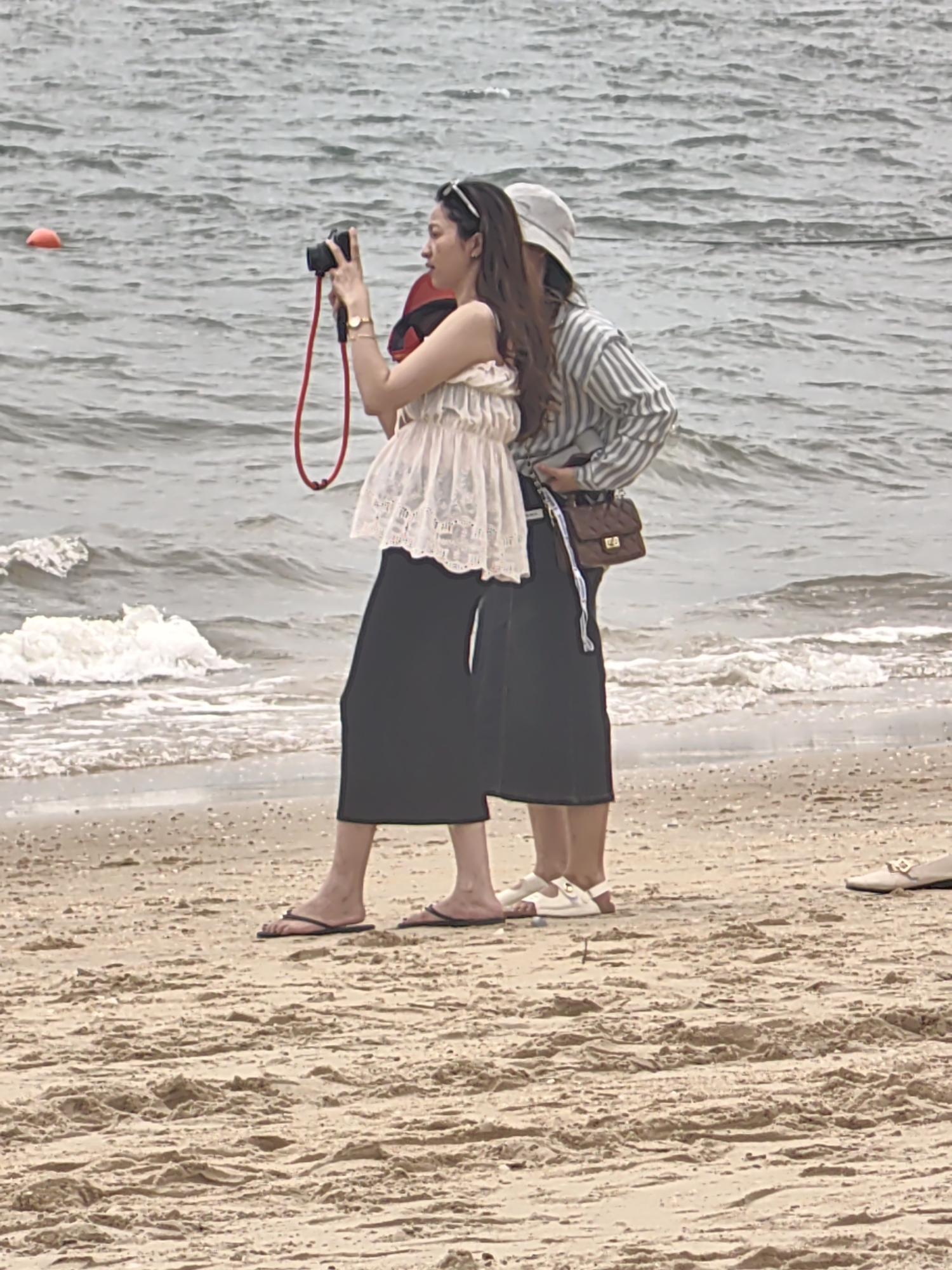 On a sandy beach in Bai Chay Ward, Vietnam, a woman stands taking a photograph with a camera that has a red strap. She wears a white, lacy top and long, dark-colored pants, and flip-flops. Another person stands behind her, wearing a striped shirt, dark pants, a light-colored hat, a brown purse, and white shoes. Waves are visible in the background. A red buoy floats in the water in the background. Another shoe is visible on the beach to the right.