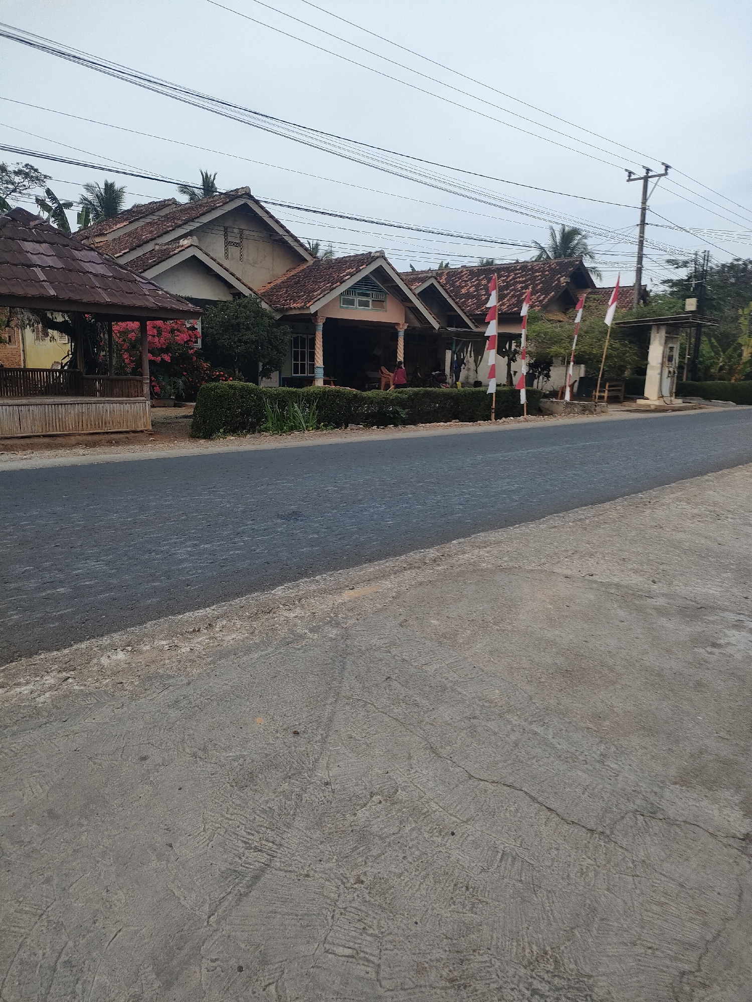 The image depicts a street scene in Sukoyoso, Indonesia. In the background, a row of traditional houses with red tiled roofs are visible. Some of the houses have porches, and one appears to be a small gas station with a pump. In front of the houses, red and white Indonesian flags are displayed on poles. A paved road runs through the foreground, with a concrete shoulder on the right. Power lines stretch across the sky above the houses. The sky is overcast. The overall impression is a quiet, rural or semi-rural residential area.