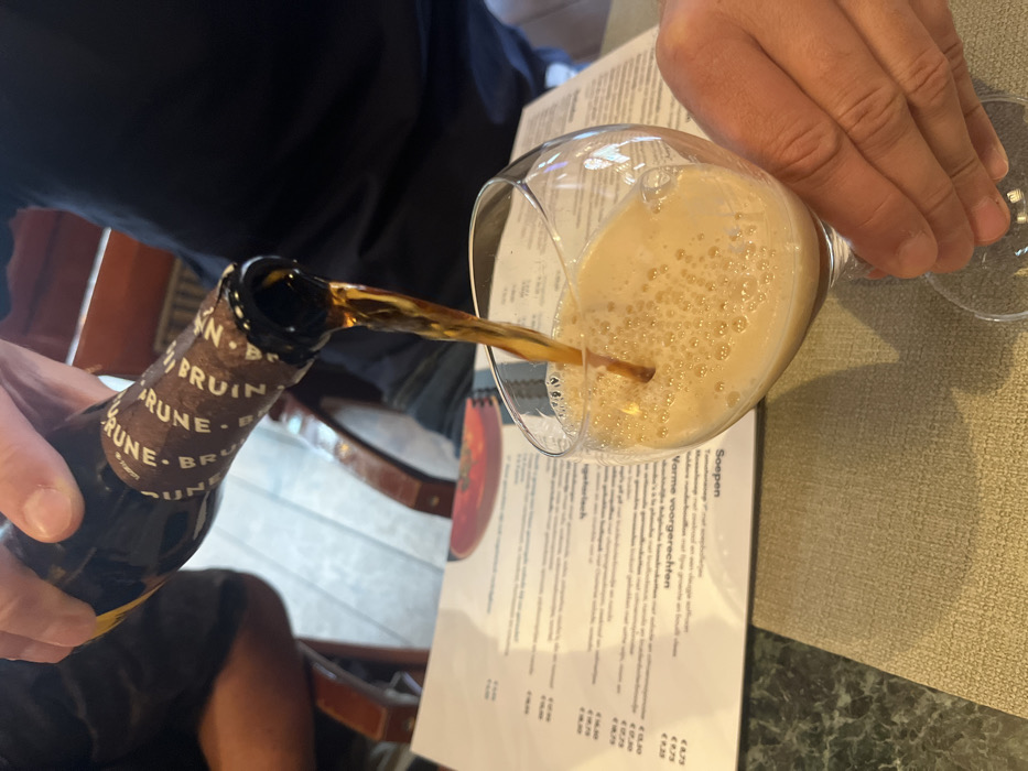 In an indoor commercial setting located in Baarle-Nassau, Netherlands, a close-up perspective captures a person's hands pouring a dark amber liquid from a brown glass bottle into a stemmed glass. The bottle's label features repeated text, partially visible as "BRUIN". The liquid creates a thick layer of tan foam within the tilted glass, which is held by the same individual. On the table, a beige woven placemat and a white menu with Dutch text, including "Soepen" (soups) and "Warme voorgerechten" (warm appetizers) alongside pricing, are present. Background elements include a dark-clothed figure and blurred wooden furniture.