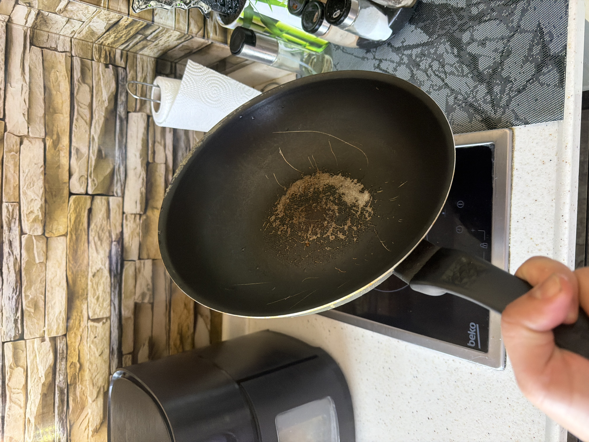 A close-up, high-angle shot captures a kitchen scene, focusing on a black frying pan or wok held by a person's hand. The interior of the pan is visibly dirty, containing dark burnt residue and several long, thin, light-colored strands that resemble human hair. The pan is positioned directly above a black induction cooktop, which features the brand name "beko" in white lettering and includes small control symbols, such as a power icon. The cooktop is bordered by a silver-colored trim and is set into a white or light gray speckled countertop.
To the left of the cooktop, a dark, partially visible appliance, possibly a bread maker or an air fryer, rests on the countertop. The background wall is adorned with a textured, faux stone or brick wallpaper, displaying various shades of light brown and beige. To the upper left, a roll of white paper towels hangs from a metallic dispenser attached to this wall. Further back on the countertop, behind the pan, a black, mesh-patterned mat or trivet is visible. Above the paper towels and behind the pan, a spice rack holds several glass shakers with black and silver caps, one of which has a green label. The overall impression is that of a domestic kitchen in Shumen, Bulgaria, capturing a moment either during or after a cooking activity, with the condition of the pan being a notable detail.