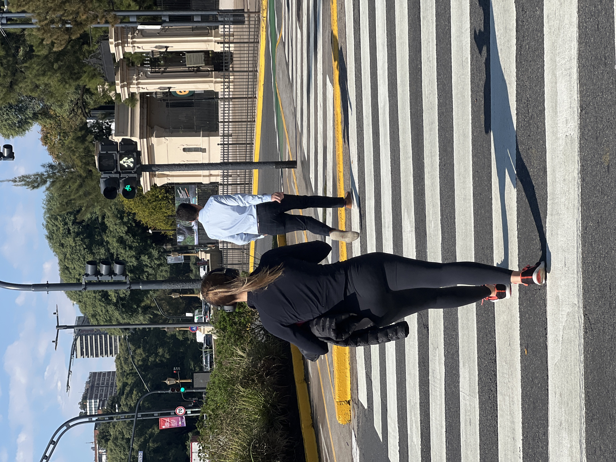 The image depicts a crosswalk in Buenos Aires, Argentina. Two individuals are crossing the street. The first, closer to the camera, is a woman wearing black leggings, a black jacket, and headphones. She also carries what appears to be a dark-colored coat. Her sneakers are visible and appear to be primarily red. The second person is a man wearing a light-colored button-down shirt and dark pants. He is walking ahead of the woman. The crosswalk consists of white stripes on asphalt, with yellow painted curbs. Traffic lights are visible, showing a green light for pedestrians. In the background, there are trees, buildings, and the sky. The presence of the pedestrian signal showing a "walk" signal indicates that the pedestrians are legally crossing the street.