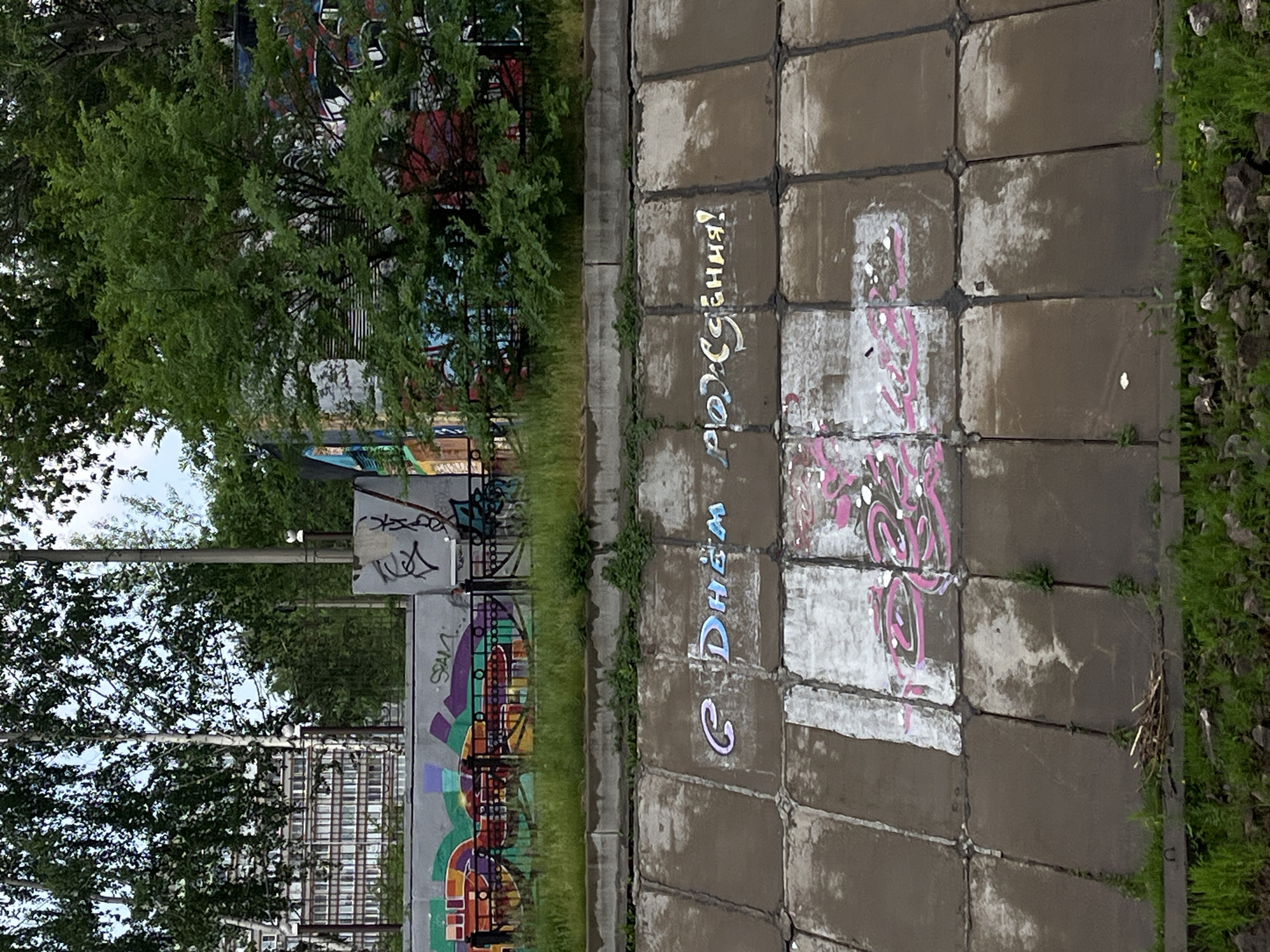 The image depicts a section of a concrete embankment lined with large, square concrete slabs, likely part of a flood control system or canal in the city of Krasnoyarsk, Russia. The embankment is adorned with graffiti. Prominently displayed in light blue and purple spray paint are the words "С Днём рождения!", which translates to "Happy Birthday!". Below this inscription, further graffiti in pink and white can be observed, with some illegible characters. In the background, a low concrete wall with colorful graffiti artwork is visible, and beyond that, a building and trees are present. The environment appears to be outdoors, with green grass growing along the bottom edge of the embankment and at the top. The overall scene is a utilitarian urban landscape altered by artistic expression.