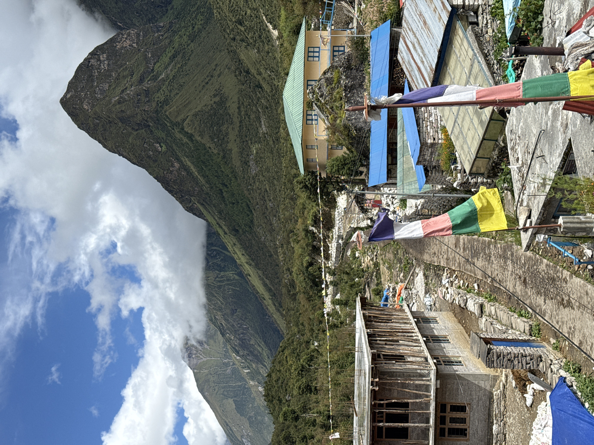 The image depicts a view of the village of Samagaon, Nepal, with a focus on its architecture and mountainous backdrop. In the foreground, prayer flags with vibrant colors – blue, white, red, green, and yellow – are prominently displayed, suggesting a spiritual or cultural setting. Several buildings are visible, including a partially constructed concrete and timber structure on the left, and a distinct yellow building with a green roof on a rocky outcrop in the mid-ground. Other structures with blue and corrugated metal roofs are also present. The environment is characterized by steep, green mountains that dominate the background, partially shrouded by clouds. A pathway or road runs through the scene, bordered by stone walls. The overall atmosphere appears to be that of a remote mountain village, likely at a high altitude, with signs of both traditional construction and ongoing development.