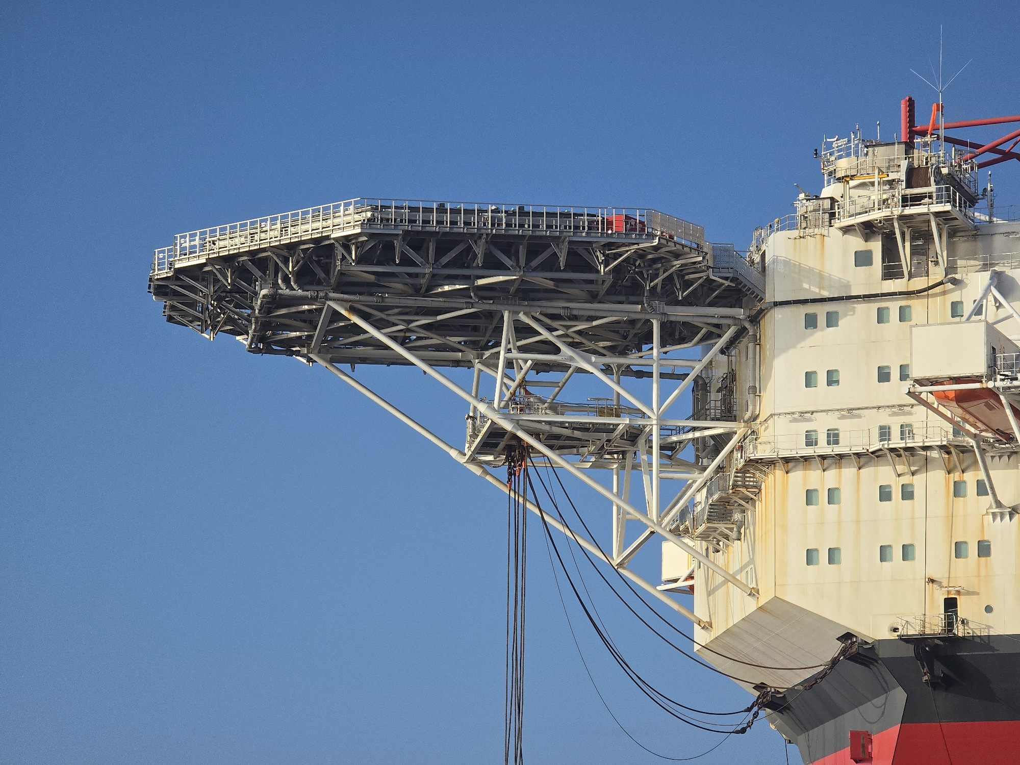 The image shows a close-up of a section of a large ship, possibly a drilling ship or crane vessel, against a clear blue sky. A helipad, constructed of a metal framework, extends outward from the side of the ship. Cables hang from underneath the helipad structure. The ship itself has a multi-story white facade with numerous rectangular windows. Rust stains streak the white paint. The lower section of the ship is painted red and black. The location and city are unknown.