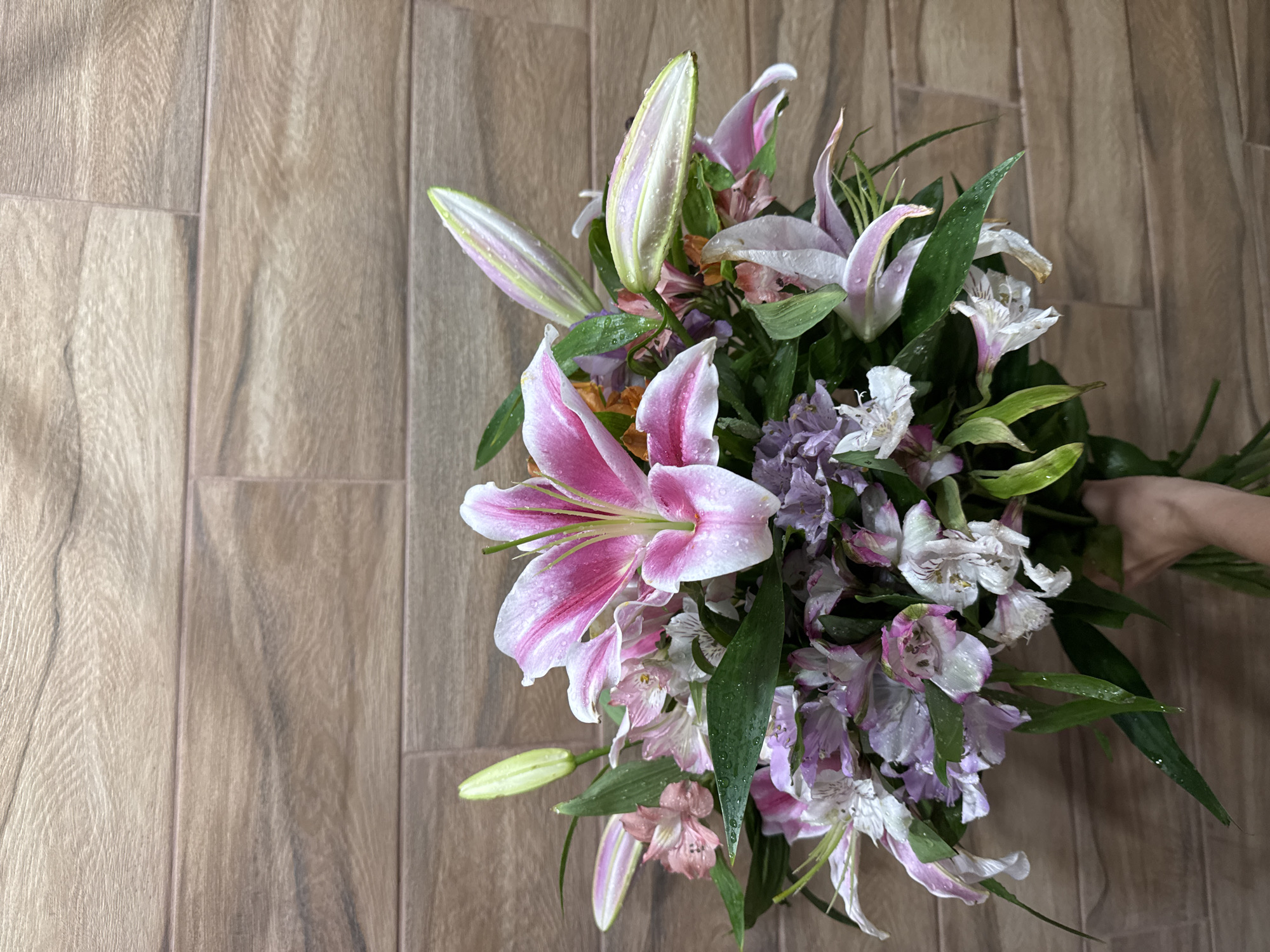 A high-angle shot shows a bouquet of flowers on a wooden surface. The bouquet features prominent pink and white lilies with water droplets, interspersed with alstroemeria in shades of white, pink, and purple. Green foliage is also visible throughout the bouquet. A hand can be seen holding the bouquet. The wooden surface underneath appears to be a floor with a textured pattern resembling wood grain.