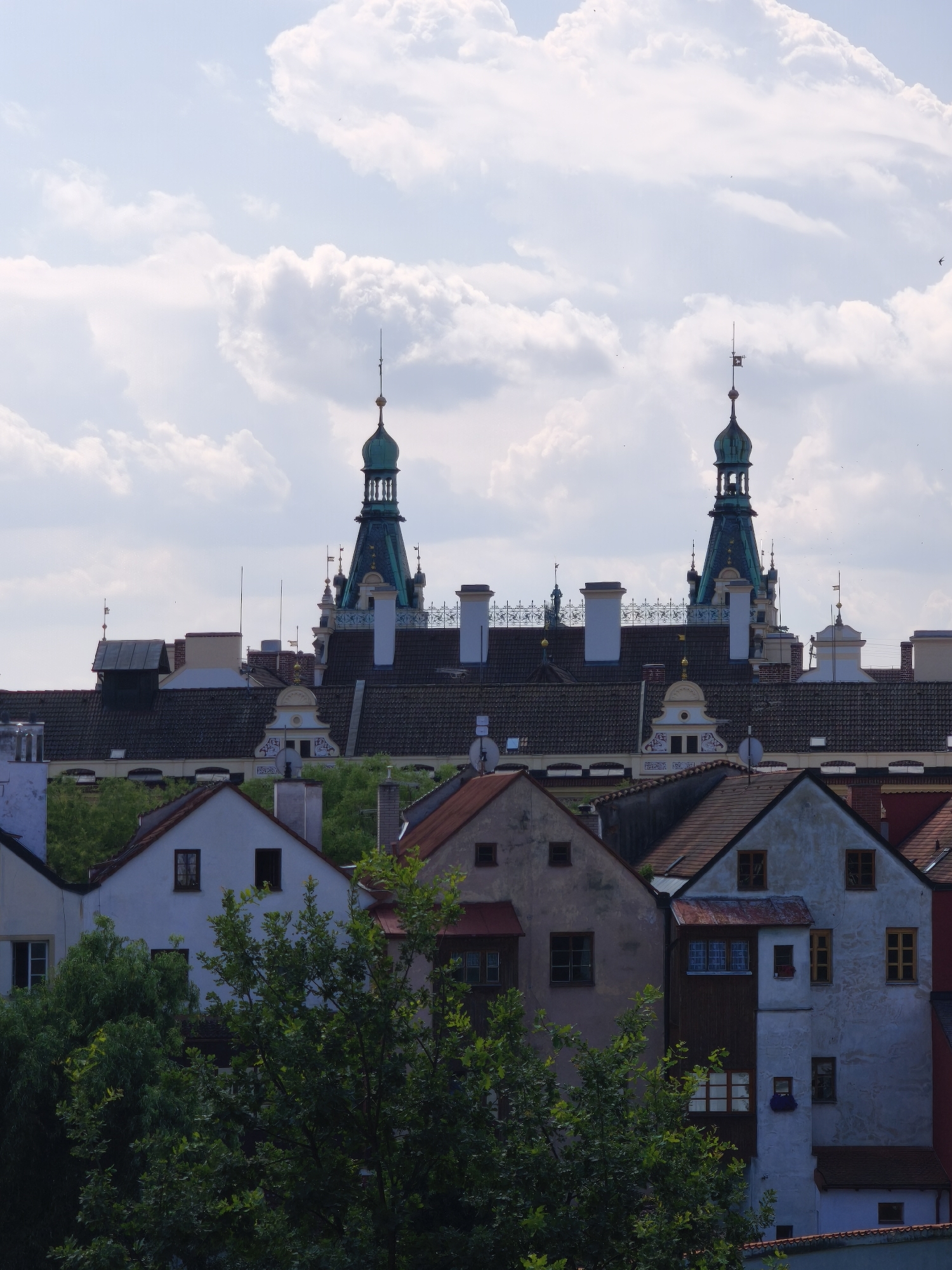 The image shows a panorama of historic buildings in Pardubice, Czechia, with a clear sky and scattered clouds. In the background, two prominent green-domed towers with ornate spires rise above a cluster of dark-tiled roofs and white chimneys. The towers are a distinctive feature of the Pardubice Castle complex. In the foreground, a row of colorful, multi-story houses with tiled roofs is visible. The houses vary in color and architectural style, with some featuring white or pastel-colored facades and wooden window frames. Lush green trees partially obscure the lower parts of the foreground buildings, suggesting a vibrant natural setting surrounding the urban architecture. The scene is captured from a slightly elevated perspective, offering a view of the city's rooftops and skyline.