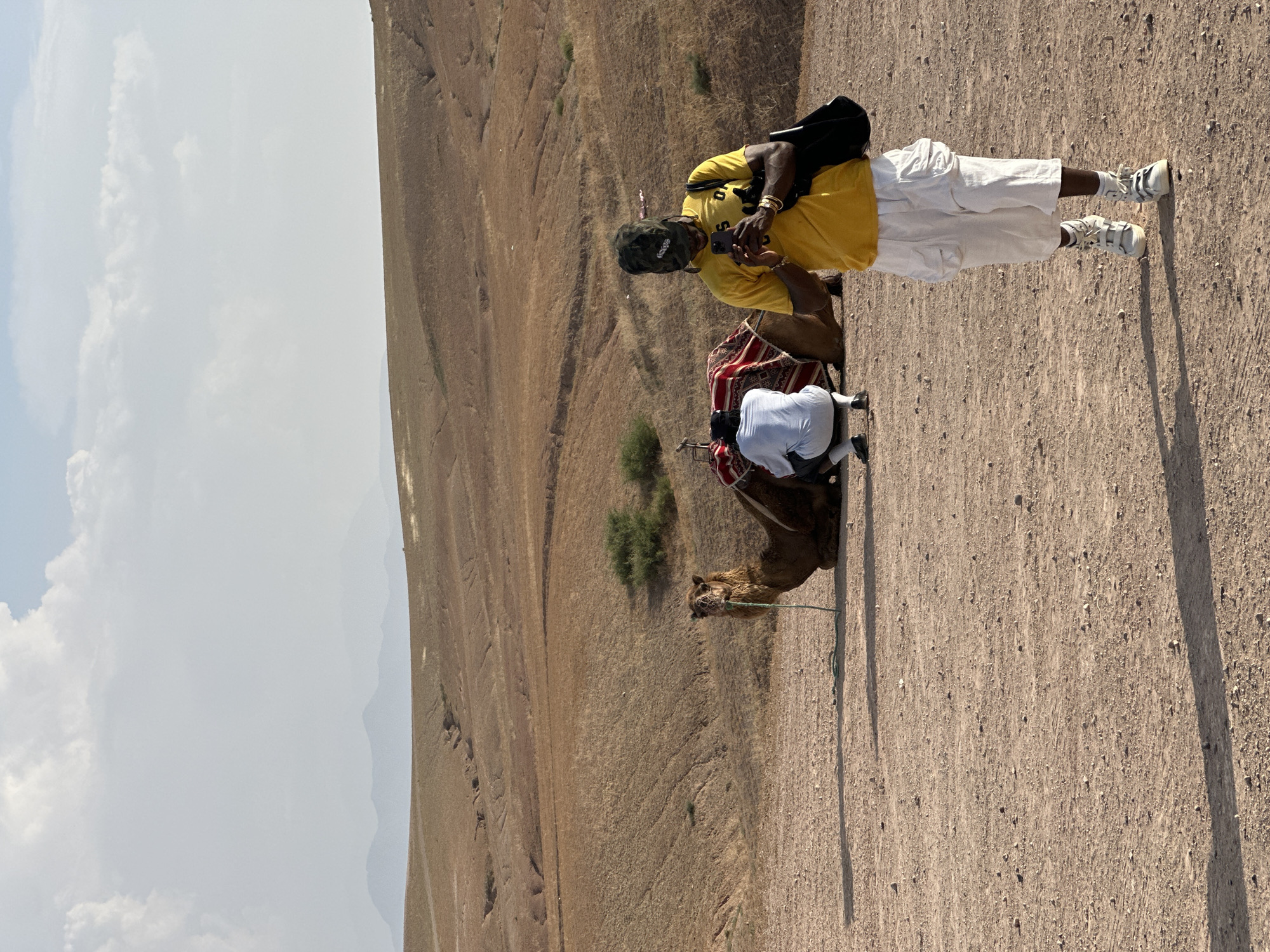 The image captures a scene in Sidi Badhaj, Morocco. A man wearing a yellow shirt, white pants, and sneakers stands near a camel. He has a black bag and is looking at a mobile phone in his hands. Another man, dressed in white, is kneeling by the camel. The camel has a saddle covered with a red and white pattern. The ground is sandy, and the background shows a dry, hilly landscape under a partly cloudy sky.