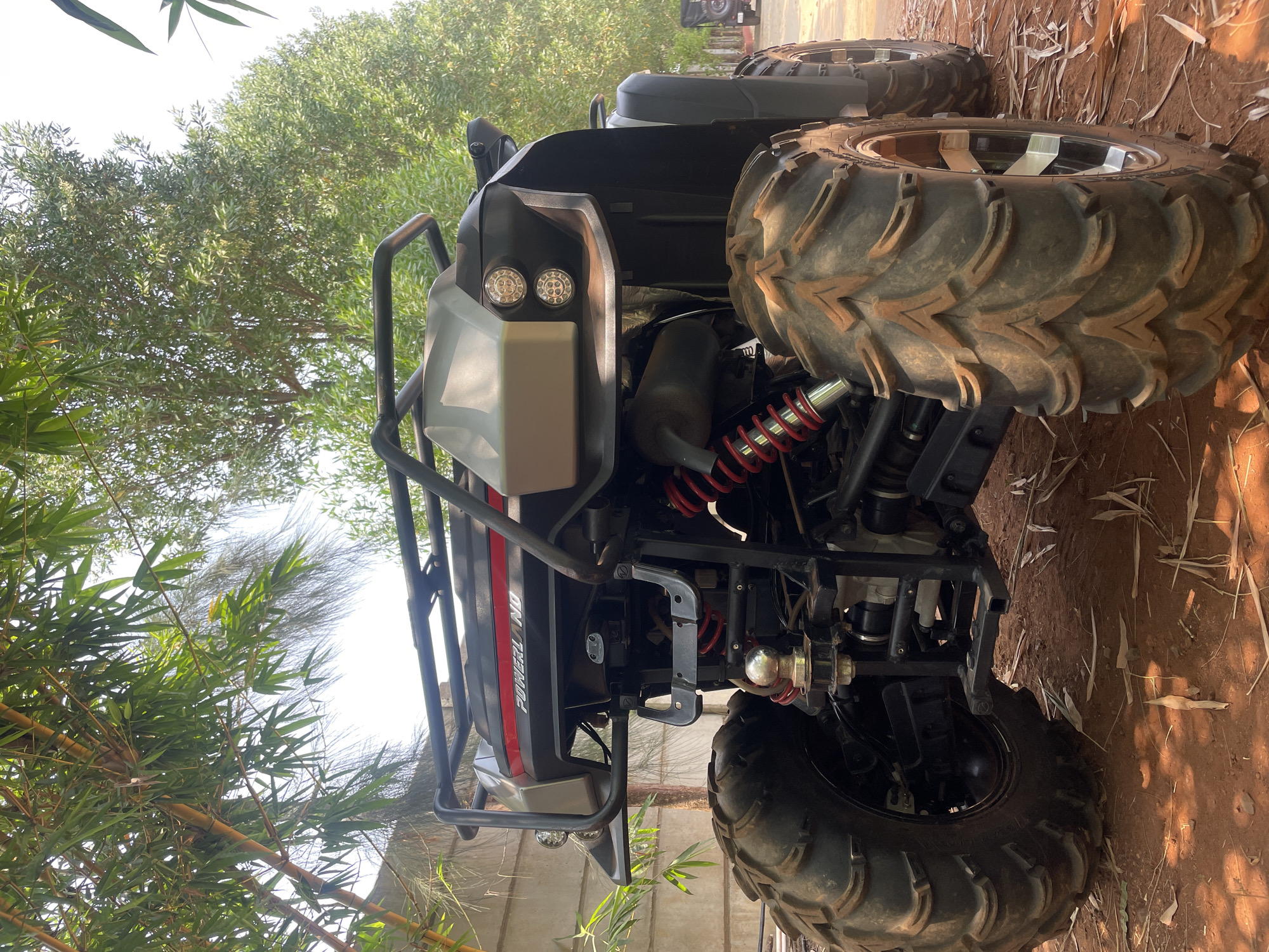 The image, taken in Dhundshi, India, presents a robust, grey and black All-Terrain Vehicle (ATV) or Utility Task Vehicle (UTV) parked outdoors. The vehicle is oriented vertically, suggesting the photo was taken with the camera rotated 90 degrees clockwise.
The ATV features large, aggressive-tread tires designed for off-road use, fitted with bright, possibly chrome or silver-colored rims. Visible components include red coil springs on the suspension, suggesting heavy-duty shock absorption, and a black exhaust pipe. The front of the vehicle is equipped with a sturdy black roll cage or bumper and two round LED headlights. A tow hitch with a chrome ball is visible towards the rear underside. The side of the ATV bears the visible text "POWERLAND" in white letters with a red accent line.
The setting appears to be a natural, unpaved environment. The ground beneath the ATV is covered in reddish-brown dirt or soil, with scattered dry leaves and debris. Lush green foliage, including bamboo plants and other leafy trees, frames the vehicle, with the bright sky visible through the canopy. The clear, bright sunlight indicates it is daytime, casting subtle shadows. No people or activities are visible; the ATV is stationary.