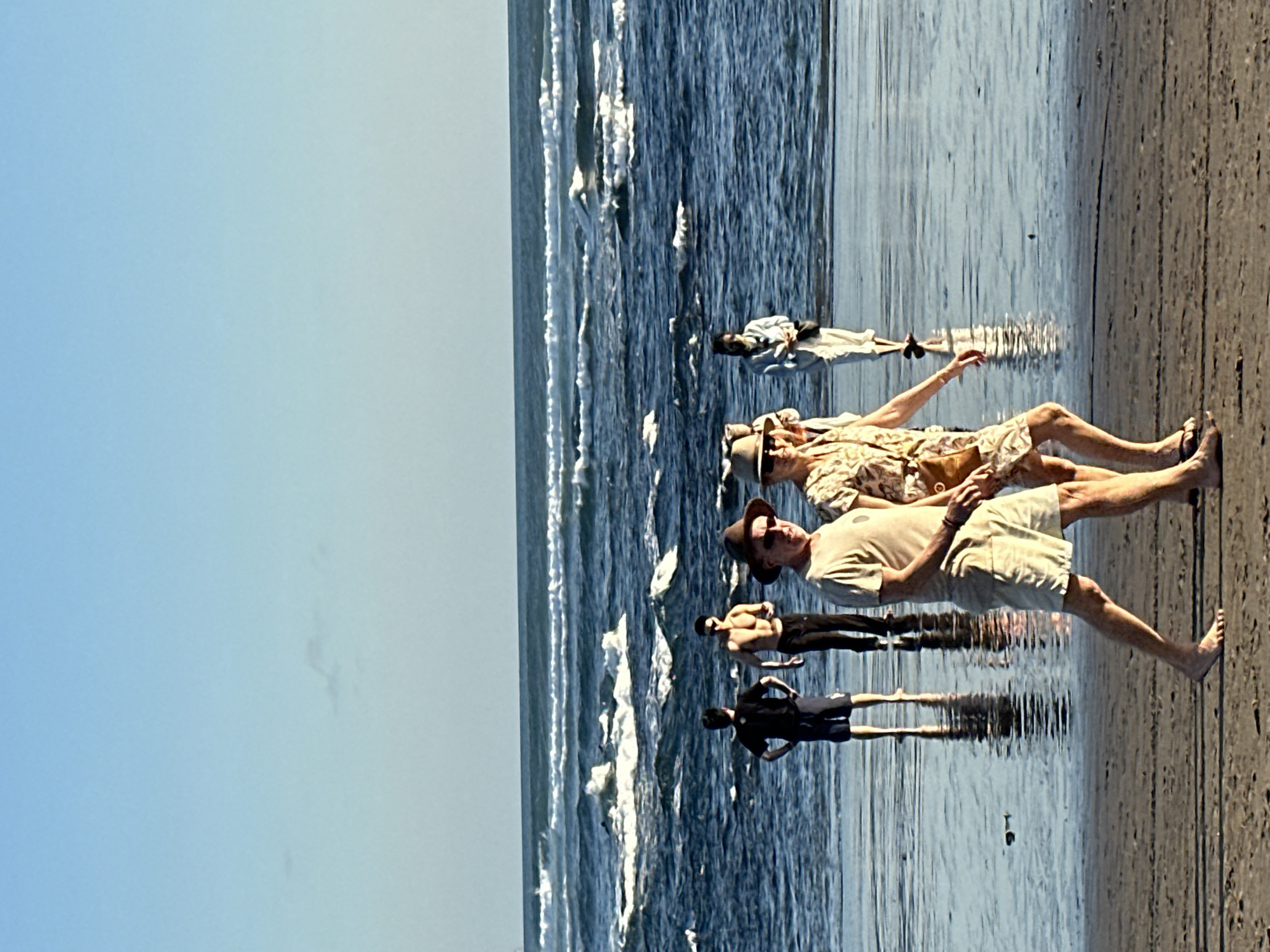 The image captures a beach scene in North Kuta, Indonesia, with several individuals present. In the foreground, a man and a woman are walking side-by-side on the wet sand. The man, wearing a light-colored t-shirt and shorts, has a hat on and is looking ahead. The woman, in a patterned dress and a hat, is also facing forward, with her hand extended slightly. Behind them, closer to the water, two men are standing, one in a black t-shirt and shorts, and the other shirtless. Further back, near the water's edge, a person in white clothing is walking away from the camera. The ocean features moderate waves with whitecaps, and the sky is a clear blue. The wet sand reflects the figures and the sky.