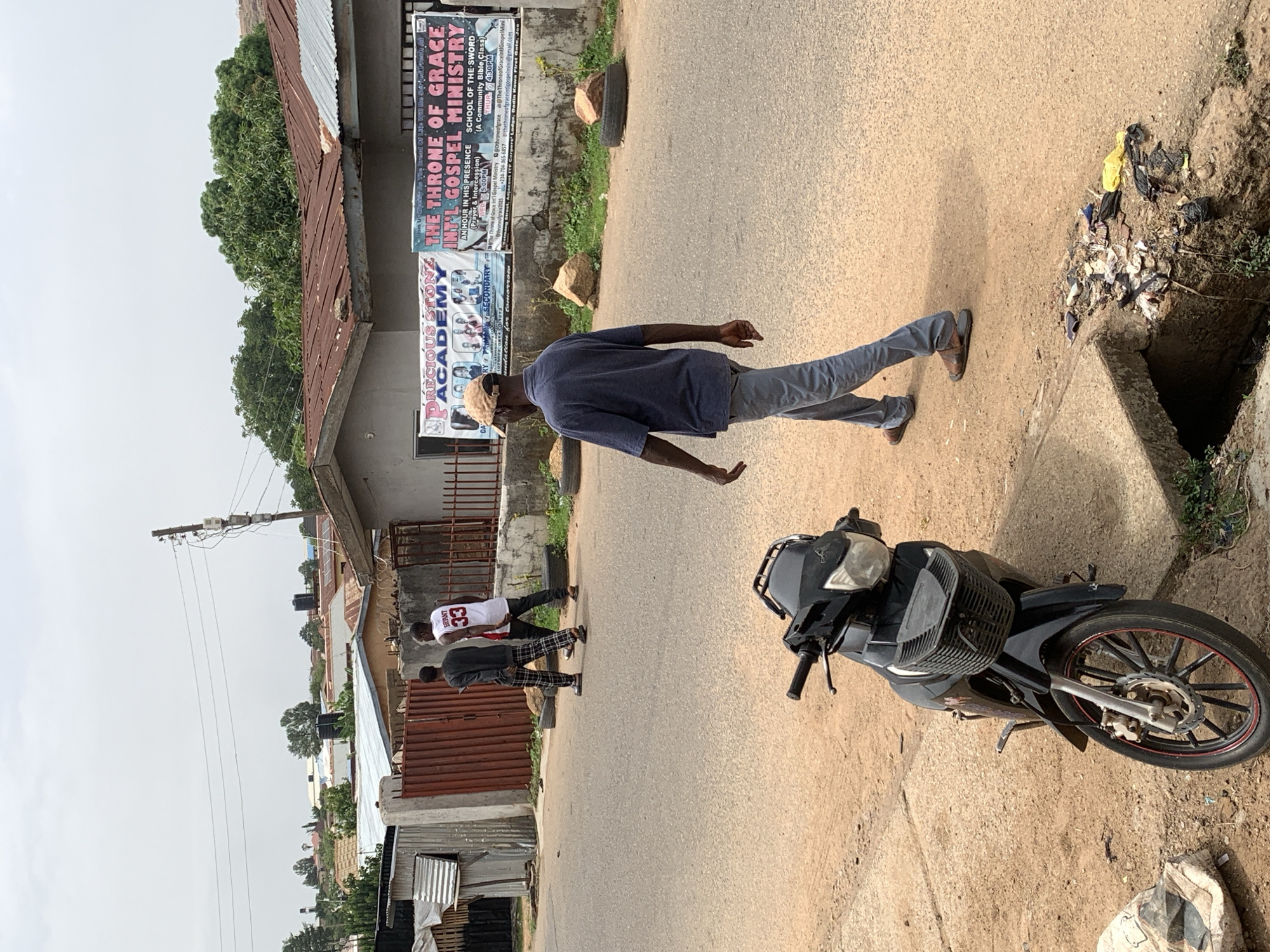 The image depicts a street scene in what appears to be the Dadin Kowa area of Jos, Nigeria, as indicated by the presence of a sign referencing "Dadin Kowa" and "The Throne of Grace Int'l Gospel Ministry." An adult male wearing a blue shirt and light-colored trousers walks along the street. Nearby, a parked black motorcycle takes up some of the road. Further down the street, two figures walk in the same direction. The figure closest to the camera wears a "33" jersey and dark trousers, while the second figure wears what appears to be a dark blazer and patterned trousers. The roadside features a storm drain filled with trash and debris. Various buildings and signs are visible along the roadside, including one for "Precious Stone Academy." Power lines are visible overhead. The overall impression is that of a residential or commercial street in a developing area.