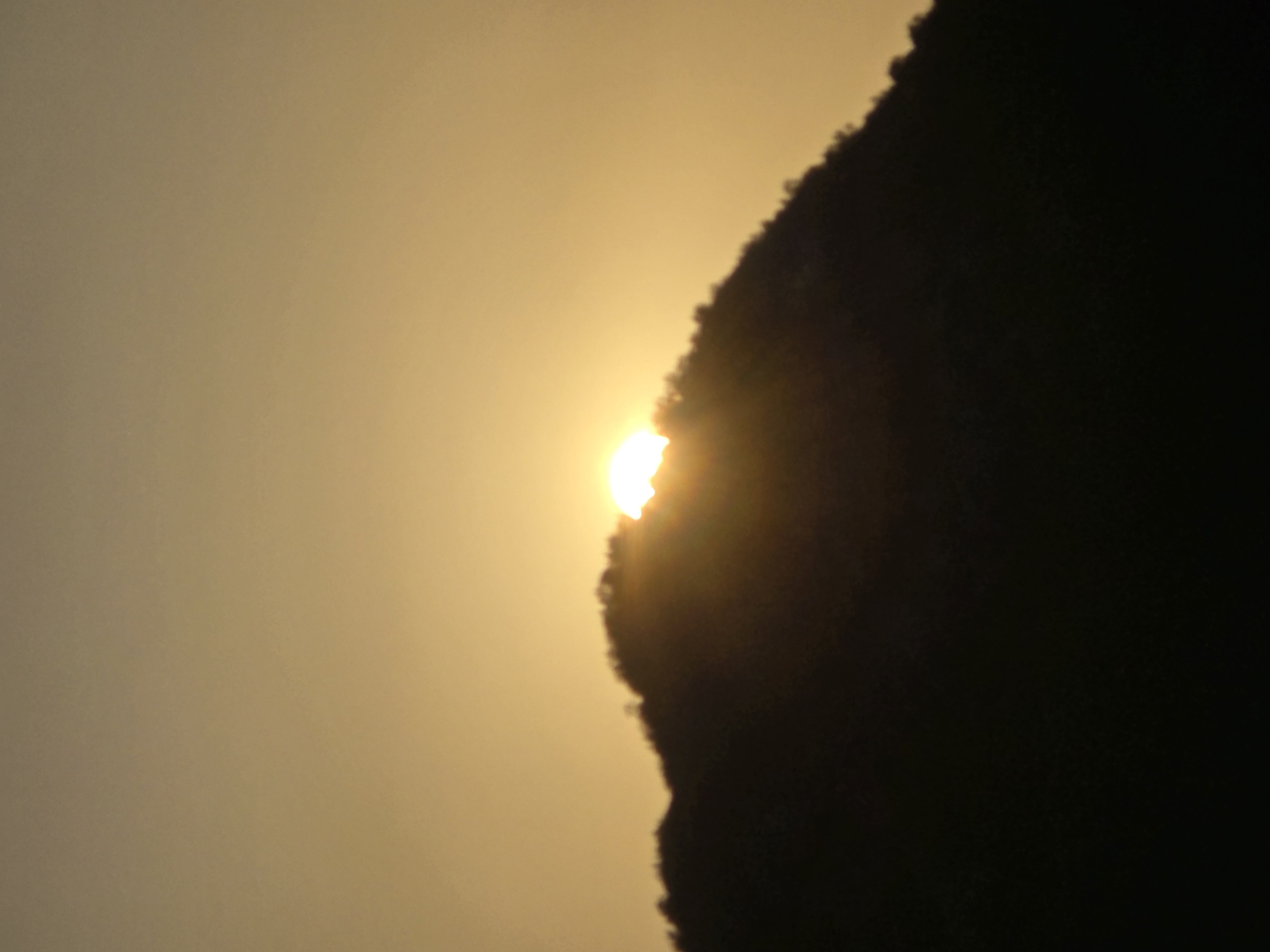 The image depicts a silhouette of a wooded mountain range against a hazy, golden sky, characteristic of a sunset or sunrise. A portion of the sun is visible, peeking over the crest of the mountain. The foreground is entirely black, obscuring any ground details. The upper portion of the image consists of a gradient of warm tones, suggesting a soft, diffused light. The photograph appears to have been taken in or near Seoca, Montenegro, as indicated by the location information. There are no people, animals, or man-made structures visible in the frame. The overall mood is serene and natural, focusing on the interplay of light and shadow on the mountainous landscape.