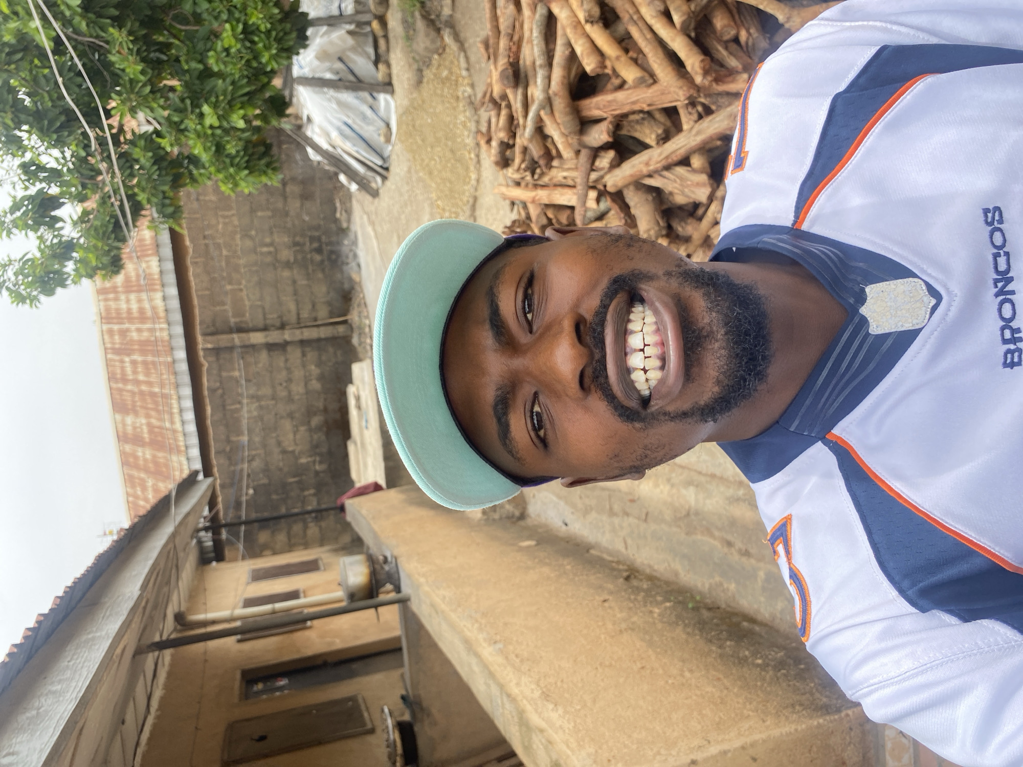 The image displays a man smiling broadly in an outdoor setting, likely in Barakin Akawo, Nigeria. The man, who has dark skin and a beard, is looking directly at the camera with a cheerful expression, revealing his teeth. He is wearing a light blue baseball cap backward and a white and blue sports jersey with orange trim. The word "BRONCOS" is visible on the right side of his jersey, and a partial number, possibly "5", is visible on the left side.
The background details suggest a semi-urban or residential area. To the man's right, there is a substantial pile of wooden logs or sticks, possibly intended for firewood or construction. To his left, a simple concrete structure, possibly a wall or counter, is visible. Further back, buildings with concrete block walls and corrugated metal roofs, some showing signs of rust and wear, are present. Lush green tree foliage is visible in the upper left of the frame. The sky is bright and overcast, indicating it is daytime. The man's wide smile conveys a sense of happiness or friendliness.