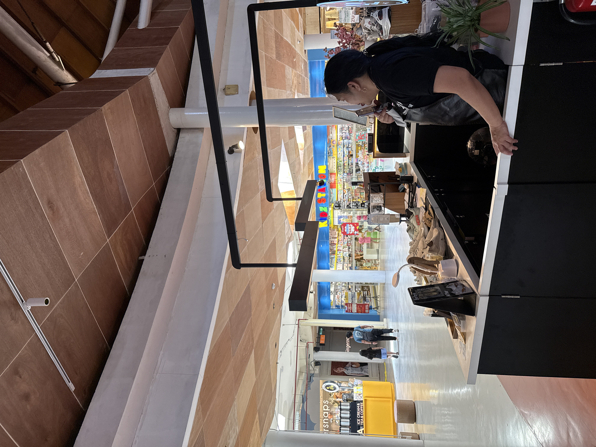 In Kuta, Indonesia, an interior scene depicts a retail environment, possibly a mall or an indoor market. The ceiling is covered with wooden panels, supported by white columns. A dark, modern counter occupies the foreground, with a woman in a black t-shirt and apron behind it, speaking on a mobile phone. On the counter are various items, including a sign that reads "Before" and "After", a small lamp, bottles, and what appear to be shoes or shoe care products. To the left of the counter, a bright yellow kiosk with the text "rsnaps" and "ICE CREAM & TEA COFFEE" is visible, with a barista or employee attending to it. In the background, two figures, a man and a child, walk away from the camera towards a bright store with colorful displays and signage, including "KIDZ STATION". Another store is visible to the far right, with merchandise and price tags. The floor is polished concrete, reflecting the ambient light.