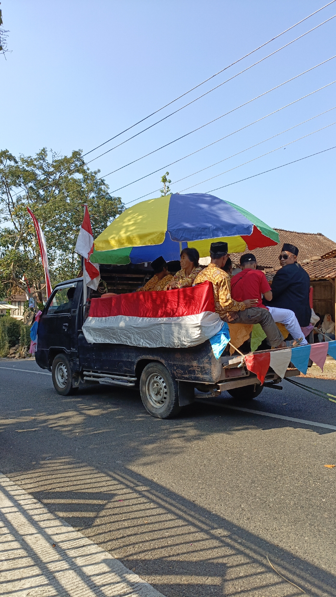 In Krajan Lor, Indonesia, on a paved road under a clear blue sky, a dark-colored pickup truck is captured in motion. The truck bed is adorned with a large Indonesian flag, its red and white bands prominently displayed. Several people are seated in the back of the truck, some under a multicolored umbrella providing shade. One individual wears a black suit and sunglasses, while others are dressed in patterned clothing and black caps. Flags and decorative pennants are also visible, suggesting a celebratory event or parade. Trees and traditional houses with tiled roofs line the background, hinting at a rural or semi-rural setting. The scene is illuminated by sunlight, casting distinct shadows from a nearby fence onto the road.