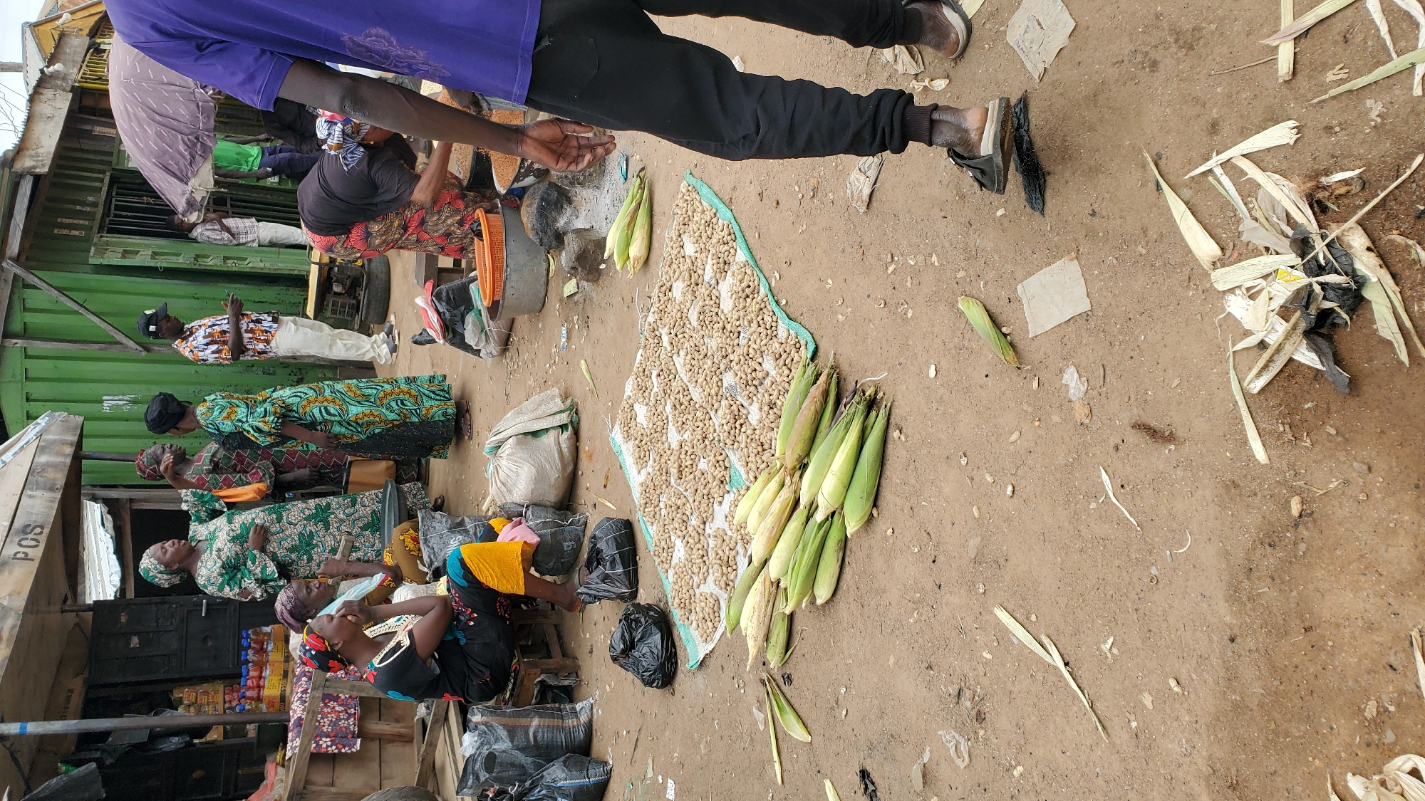 In Jalingo, Nigeria, a street market scene unfolds with several people engaged in commerce. In the foreground, a pile of corn stalks lies on the dusty ground, interspersed with discarded leaves and debris. To the right, a person wearing black pants and a purple shirt stands with their feet visible, one sandal worn. Further back, a spread of peanuts lies on a blue tarp, a common method of drying produce. Several women in traditional attire are visible, some seated and others standing, engaged in conversation or tending to their goods. Bags of produce and other items are scattered around, indicative of a bustling marketplace. The background shows market stalls and a green shipping container, suggesting a semi-permanent setup for vendors. The overall impression is one of a lively, everyday market scene in an urban Nigerian setting.
