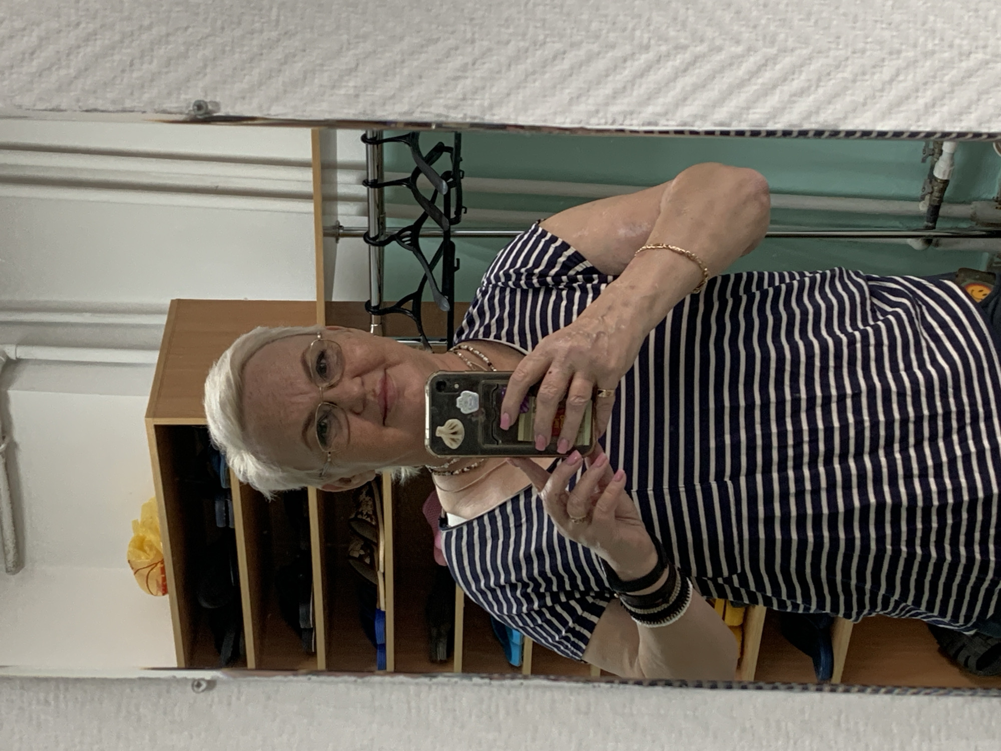 An older woman is taking a selfie in a mirror. She is wearing a navy and white striped shirt and eyeglasses. Her hair is short and gray. She has a gold bracelet on her right wrist and multiple bracelets on her left wrist. She is holding a black smartphone in her hands. In the background, there is a wooden shelving unit with various items inside, a coat rack, and a light green wall. Based on prior knowledge, this could be in Chaikovsky, Russia.