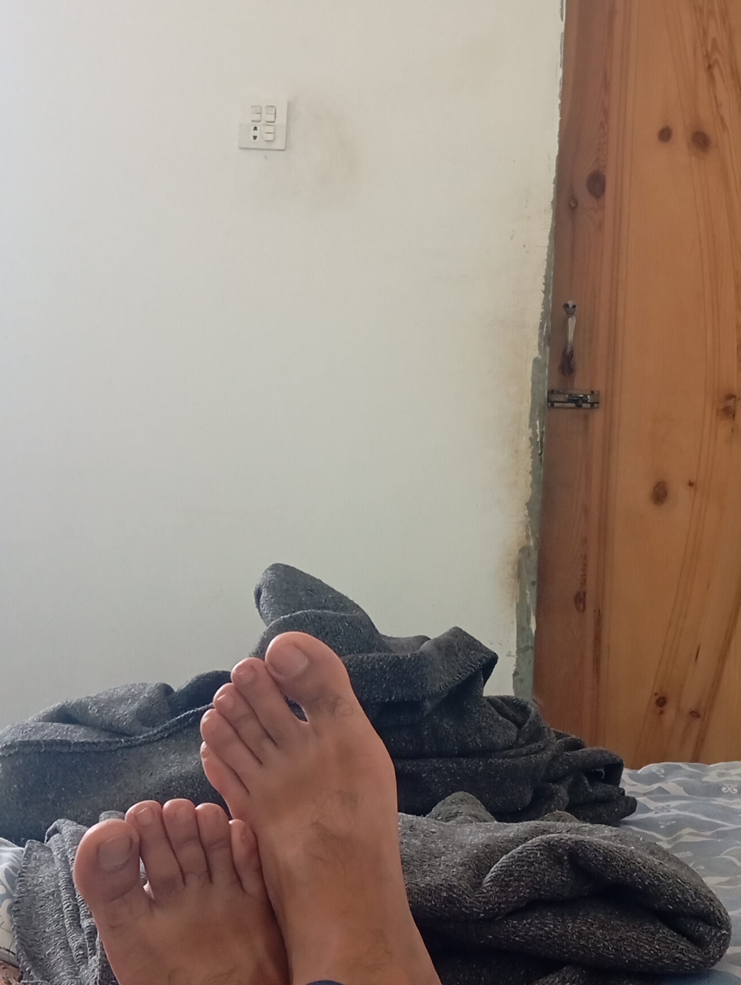 The image depicts a close-up view of two bare feet resting on a bed. The feet are positioned on top of dark grey blankets, which are draped over a patterned bedspread. The skin tone is medium to dark. The background shows a white wall with an electrical outlet and a wooden door with a simple latch. The lighting appears to be natural, casting soft shadows. The scene suggests a domestic indoor setting. No specific location in Lassan, Pakistan is evident from the visual cues.