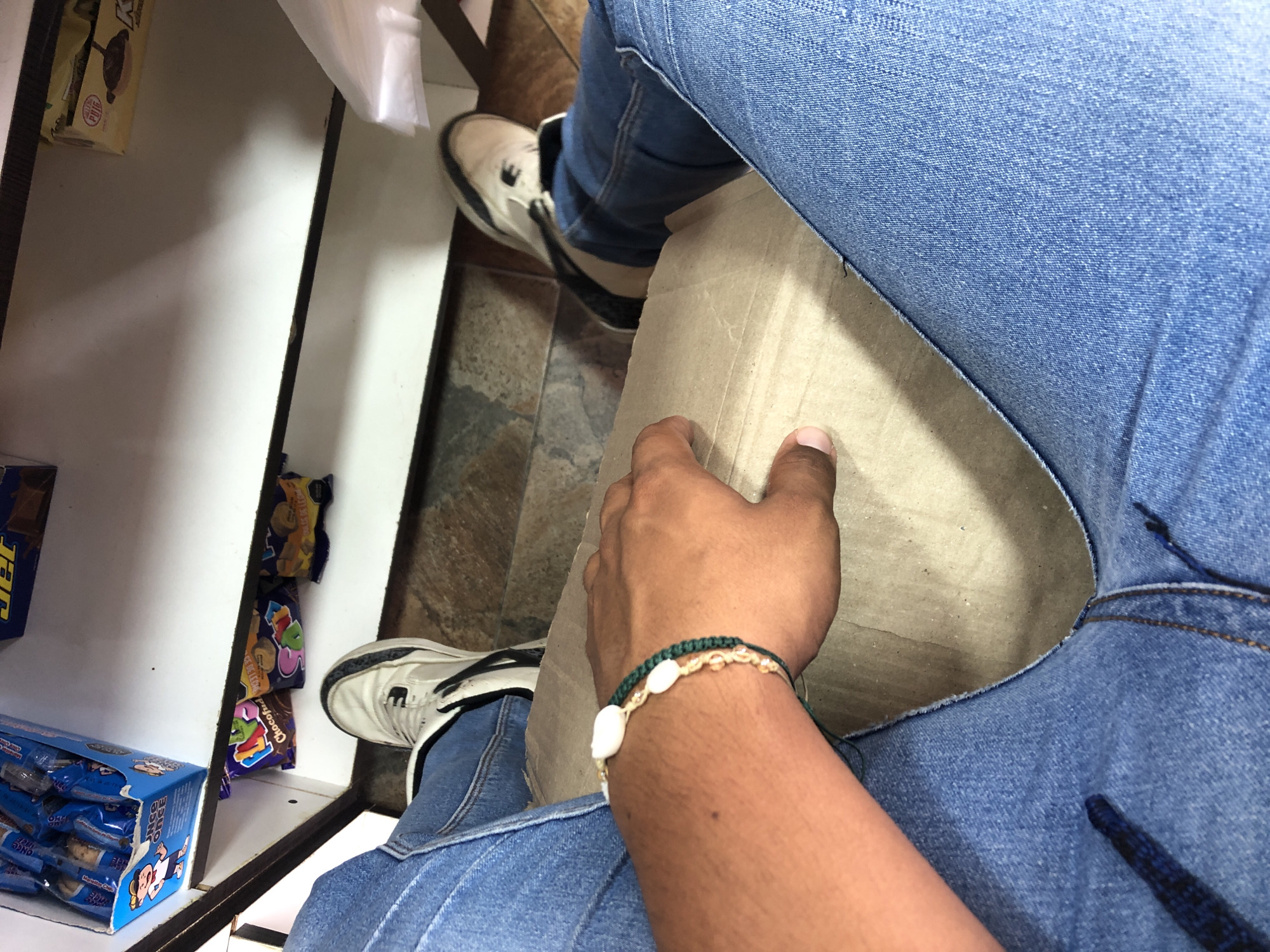 The image shows a first-person perspective of someone sitting in what appears to be a small shop or store in Juan Griego, Venezuela. The individual is wearing blue jeans and white sneakers. A cardboard box is on the person's lap. The person's right hand rests on the box, and a bracelet made of green cord and white beads or shells is visible on their wrist. To the left, shelves are stocked with various packaged snacks and candy. The floor has a tile pattern.