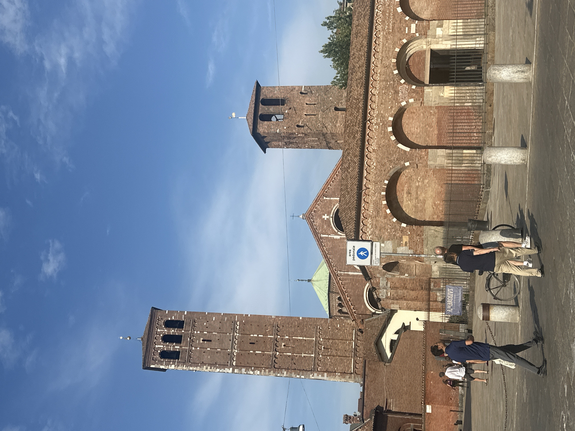The image depicts the Basilica di Sant'Ambrogio in Milan, Italy, on a clear day under a bright blue sky. The basilica features two prominent bell towers, one taller and to the left, and another shorter one to the right. The architecture is characterized by red brickwork, arched doorways, and detailed ornamentation. A green-domed structure is visible behind the main facade, and a cross adorns the roof.
In the foreground, several people are walking across a paved plaza. Two men in the center foreground are walking towards the right. The man on the left wears a dark blue shirt and grey pants, carrying a white bag. The man to his right wears a dark blue polo shirt and light-colored pants. Behind them, another man, bald and wearing a dark t-shirt and shorts, walks with a bicycle. Further to the left, two men and a woman are walking away from the camera. A sign indicating a pedestrian area ("AREA PEDONALE") with a walking figure is mounted on a pole to the right of the main entrance. A dark banner with the word "AMBROSIUS" is visible in the background. The plaza is furnished with bollards and a metal fence bordering the basilica's facade. The scene captures a moment of everyday life in front of a historic religious site.