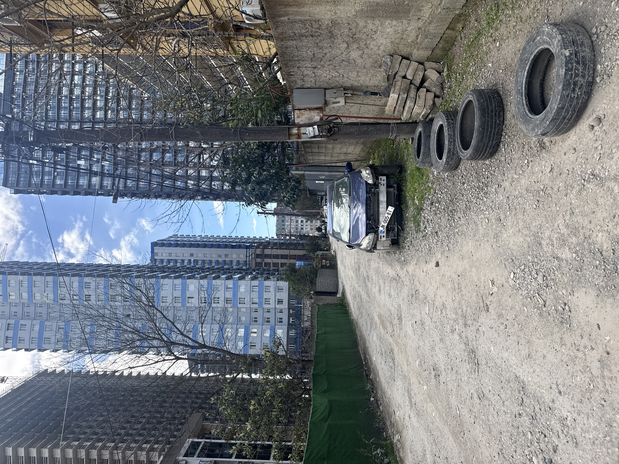 The image depicts a narrow, gravel alleyway in Batumi, Georgia. Tall, modern apartment buildings line one side, while a low concrete wall and fence define the other. An abandoned blue car, heavily damaged in the front, sits in the middle of the alley, its license plate reading "JK-005-BI." To the right of the car are four used tires and a pile of concrete blocks. A utility pole, equipped with electrical boxes and wires, stands near the car, connecting to overhead lines. The sky is partly cloudy, with patches of blue visible between the buildings. There are no people visible in the image.