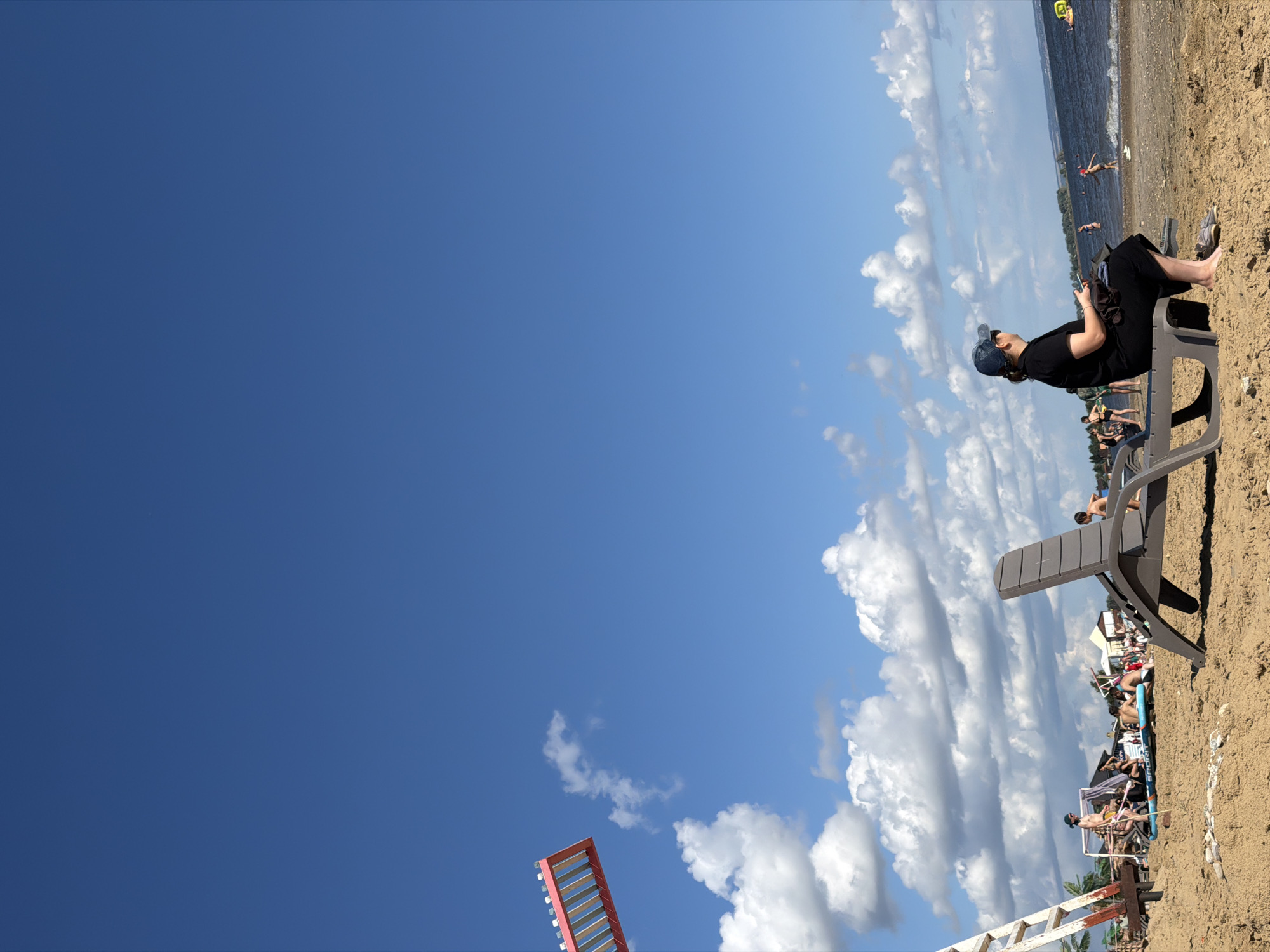 The image depicts a sunny day at a beach, likely a waterfront area within the city of Unknown. The sky is a vibrant blue with scattered white clouds. In the foreground, a person wearing a dark outfit and a blue cap is relaxing on a grey lounge chair, facing away from the camera and towards the water. To the left, a red and white structure, possibly part of a Ferris wheel or a similar attraction, is partially visible against the sky, along with a ladder. Further down the beach, several other people are visible, some wading in the water and others sitting or standing on the sand, suggesting a recreational atmosphere. The sand is golden, and the water appears to be a lake or a calm sea, with small waves lapping at the shore. The background shows a distant treeline and some structures, indicating the presence of a town or city nearby.