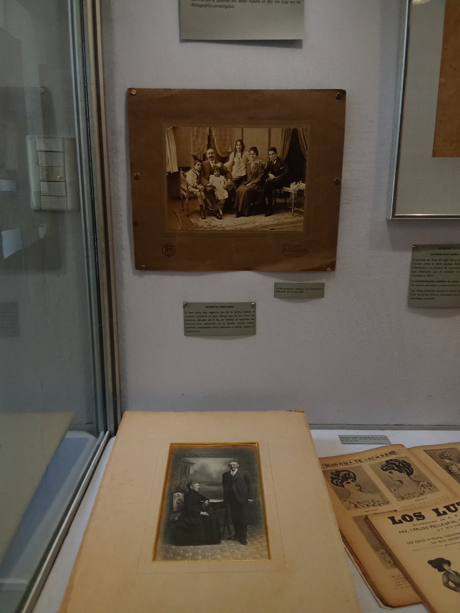 The image shows an exhibition display inside a glass case, likely at a museum or gallery, located in Buenos Aires, Argentina. The display features several framed photographs and printed materials.
In the upper part of the display, a large, sepia-toned photograph in a brown mount depicts a family posing for a portrait. The family consists of adults and children, all dressed in formal attire. The composition suggests a formal, indoor setting with draped curtains in the background.
Below this larger photograph, a smaller framed photograph shows a man and a woman seated, with a man standing behind them. The couple in the foreground is dressed in formal attire, while the standing man wears a suit and holds a cane. The background appears to be a studio setting with a painted backdrop.
To the right of the central display, several other framed items are visible, including what appears to be a tall, narrow framed print or photograph.
In the foreground, a stack of magazines or printed publications is partially visible. One of the visible titles reads "LOS LU" which is likely part of "LOS LUNES DE LA MODA," a fashion magazine. The content of these publications includes illustrations of women's hairstyles and fashion from an earlier era.
The overall lighting of the scene is consistent with museum exhibition lighting, highlighting the artifacts within the display case. The background of the exhibition space is a textured, off-white wall. Some text is visible on small plaques accompanying the photographs, providing context or descriptions of the exhibited items. One plaque reads "RETRATOS FAMILIARES" (Family Portraits).