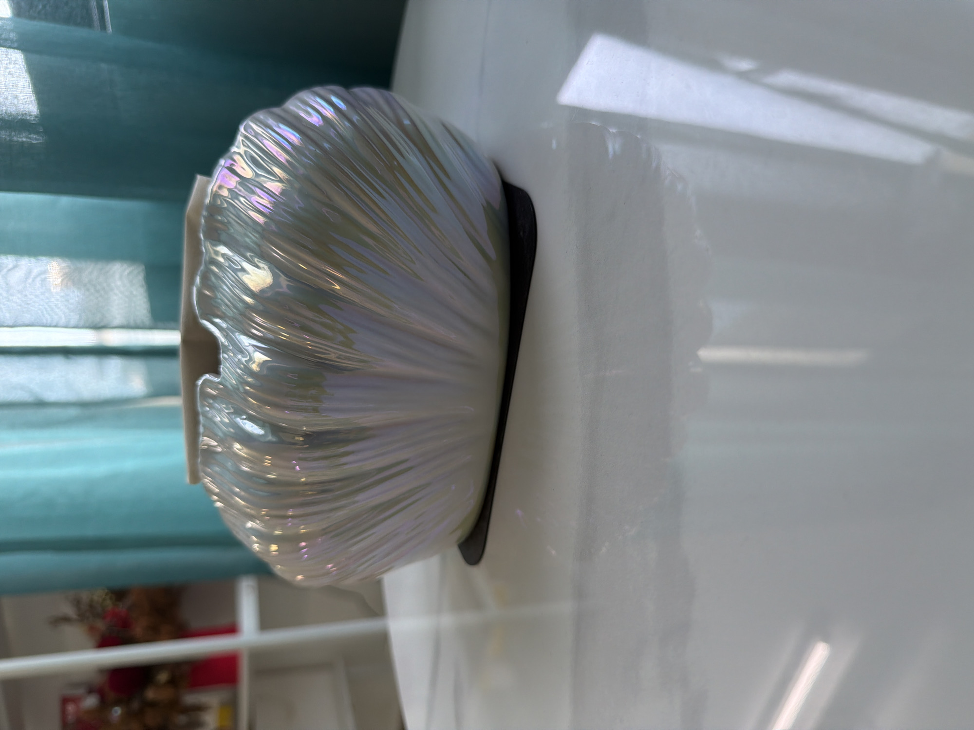 A pearlescent ceramic bowl with a fluted design sits on a white marble-patterned table. The bowl has an iridescent sheen with hints of pink, blue, and green. It rests on a small, black, flat base. In the background, a teal curtain is visible, partially obscuring a white shelf unit that holds a red vase with dried floral arrangements. The overall scene is indoors, suggesting a home or decorative display setting. No specific city or ward information is discernible from the image itself.