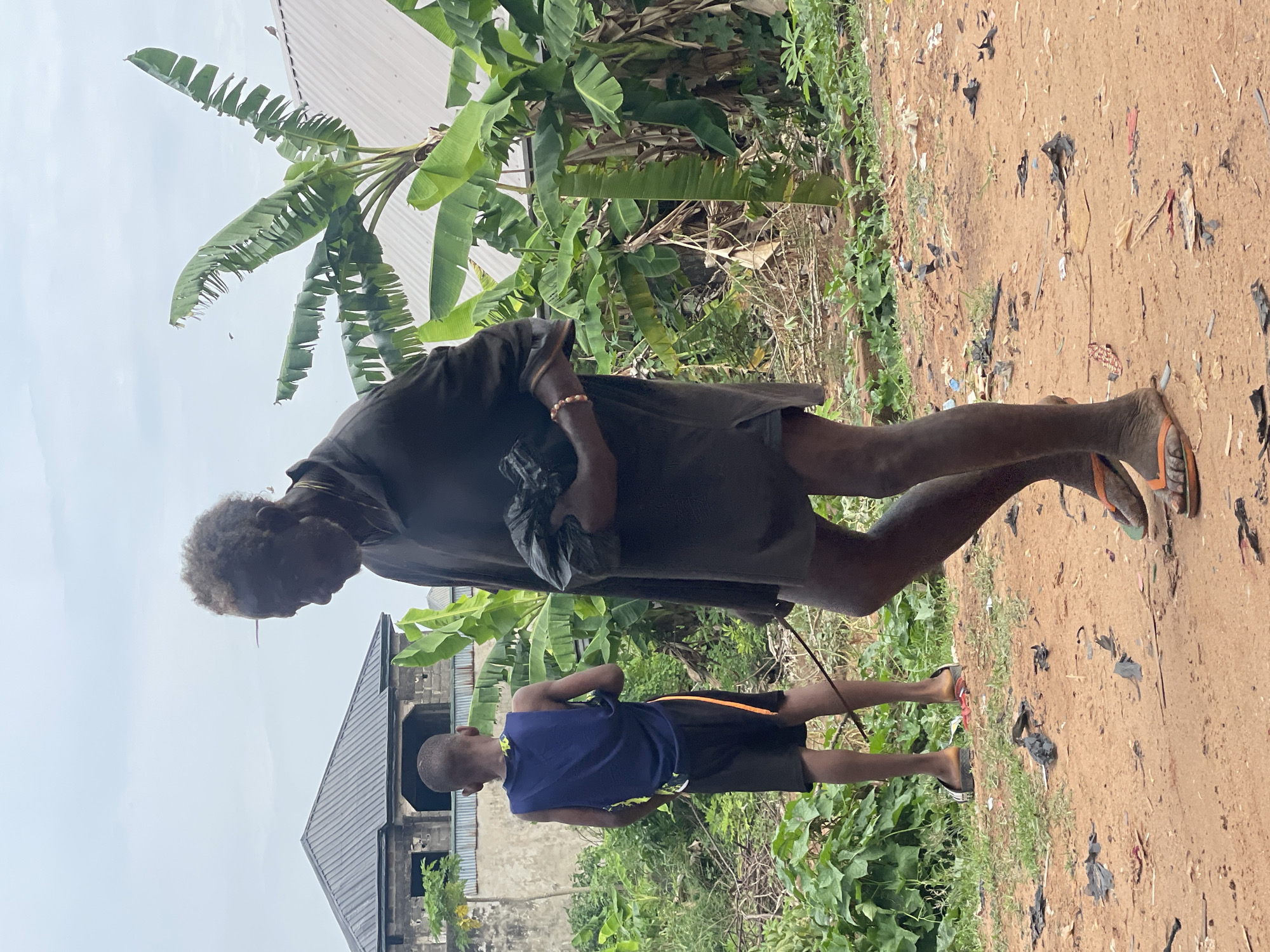 In Onitsha, Nigeria, an elderly person with dark skin and graying hair walks on a dirt path strewn with litter, wearing a dark-colored, knee-length garment and flip-flops. They are holding a black plastic bag. Beside them walks a younger person, possibly a child, wearing a blue sleeveless top and dark shorts with an orange stripe, also in flip-flops. They appear to be connected by a slender stick or cord. The surrounding environment consists of lush green vegetation, including banana trees, and a building under construction with a gray metal roof is visible in the background.