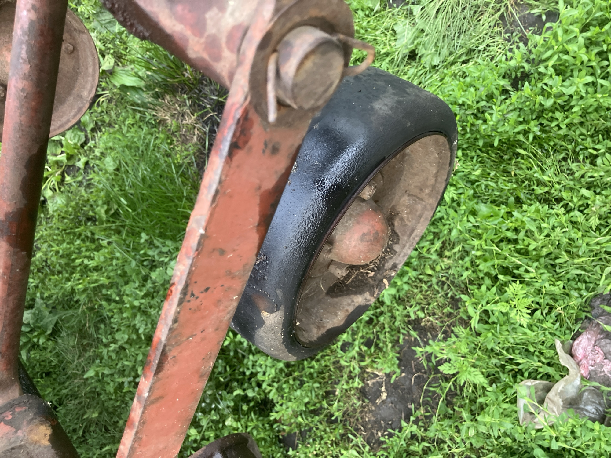The image shows a close-up, overhead view of a worn, red metal frame resting on green grass in Vovkivka, Ukraine. A flat, black tire with a reddish-brown hub is visible, attached to the metal frame. The tire appears to be covered in a thin layer of moisture. The grass is dense and uneven, with some patches of bare earth. In the lower left corner, a crumpled piece of light-colored plastic or fabric is visible amongst the grass. The overall scene suggests an outdoor environment that is likely unkempt or rural.