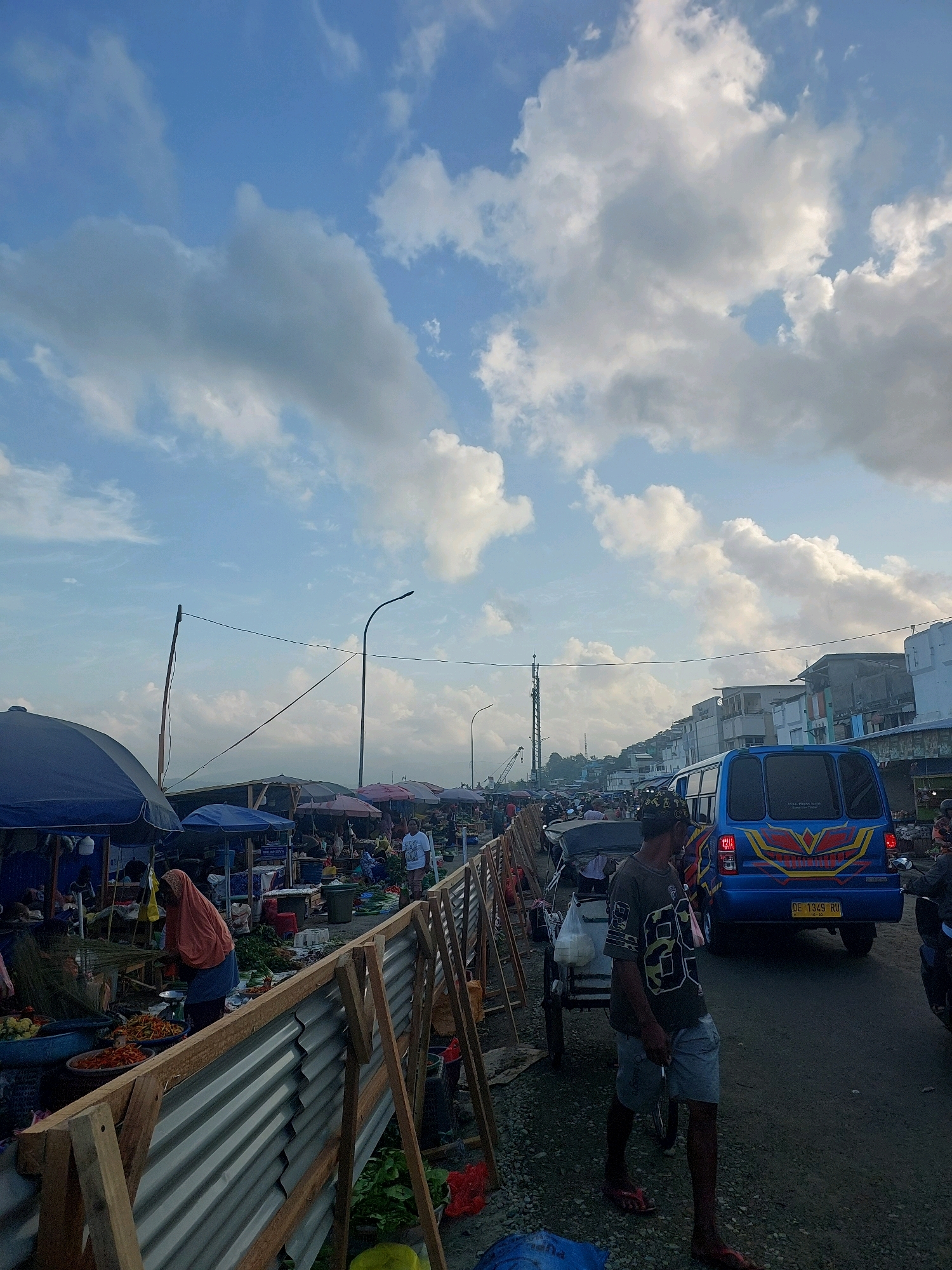 The image depicts a bustling outdoor market in Ambon, Indonesia, likely a roadside market given its proximity to a street with vehicles. The sky is a bright blue, dotted with fluffy white clouds, suggesting it's daytime, possibly late afternoon due to the soft lighting. In the foreground, a temporary barrier made of corrugated metal and wooden frames separates the market stalls from the pathway.
Several market stalls are visible, covered by blue and dark blue umbrellas, selling various goods, predominantly fresh produce like chilies and leafy vegetables. People are present, some engaged in selling and others browsing or walking through the market. One woman in an orange headscarf and blue skirt is seen attending to the produce at a stall. Further down the market, more stalls and people are visible, extending into the distance.
On the street, a bright blue van with red and yellow accents is prominent, with a license plate visible. Several other vehicles, including motorcycles and cars, are present on the road, indicating the market's location alongside a busy thoroughfare. A man in a dark t-shirt and shorts is walking in the foreground, carrying something in his hand. Another man, wearing a cap with a distinctive pattern, is also visible near the blue van. The overall scene conveys a lively atmosphere of daily commerce in a tropical urban setting.
