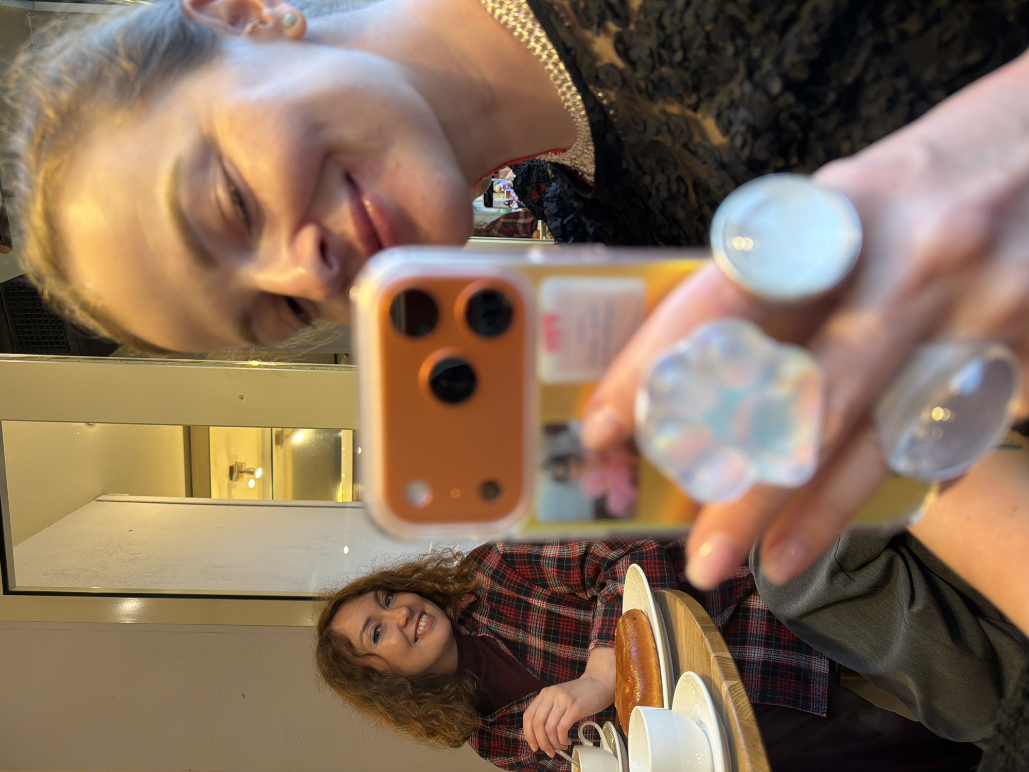 A woman takes a selfie in a mirror. The woman's reflection is of a person seated at a table with a plate containing a pastry, and cups/saucers on the table. The person in the reflection has curly hair and is wearing a red and black plaid shirt. The location cannot be determined from the visual information.