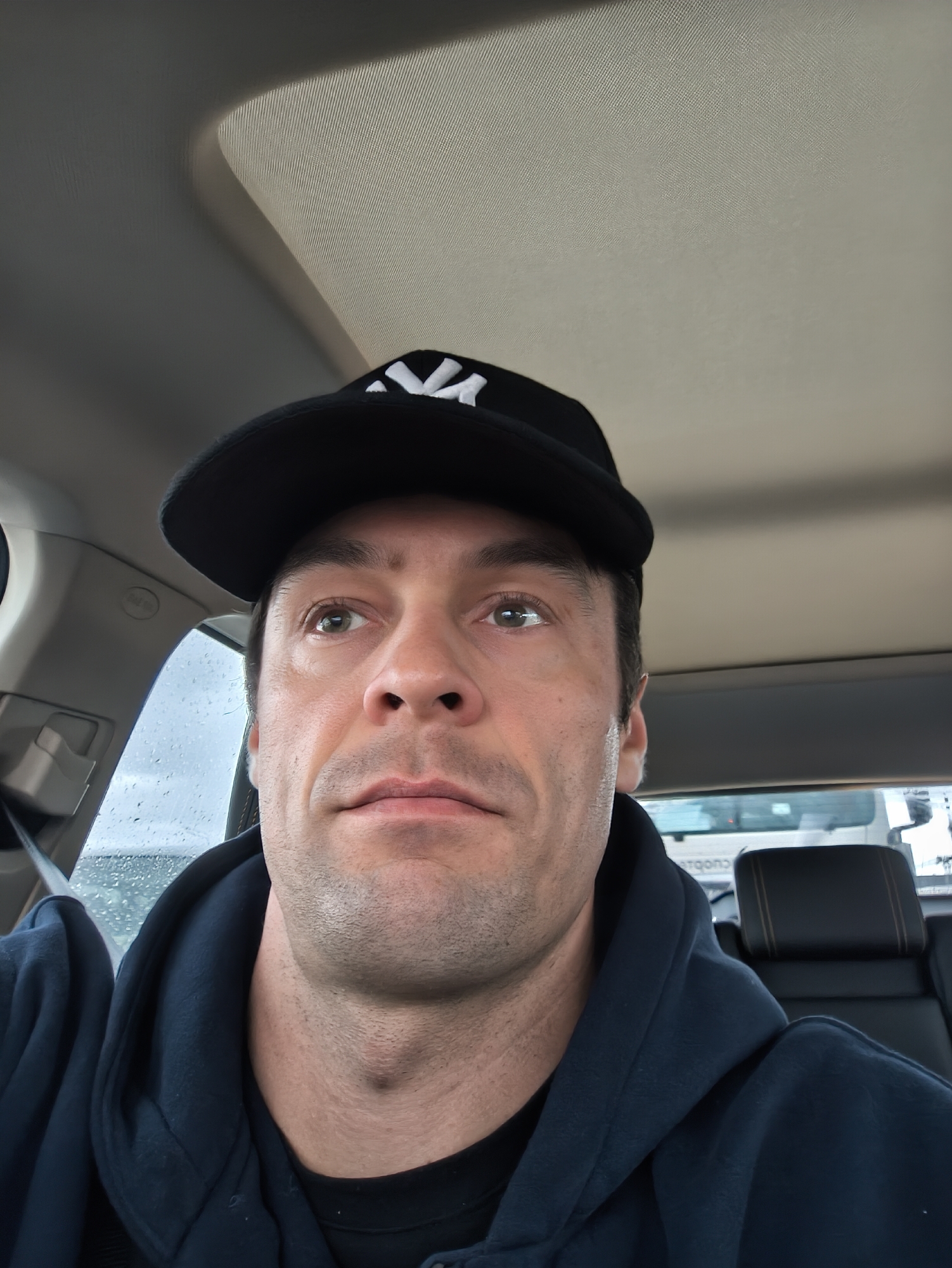 A man is in the driver's seat of a car. He is wearing a black baseball cap with a white logo. He is also wearing a dark blue hooded sweatshirt. The interior of the car has a light-colored roof. Rain is visible on the side window. The location is not discernable as Moscow, Russia, from the visual information provided.