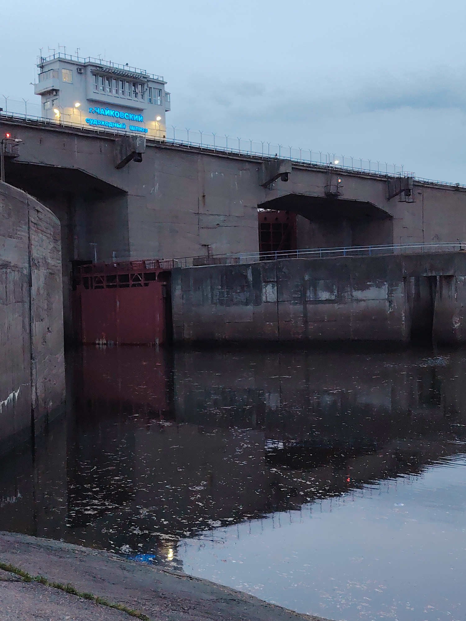 This image depicts a section of the Chaikovsky Shipping Lock in Chaikovsky, Russia. The lock is constructed of concrete with a red metal gate visible on the left side of the frame. The water in front of the lock is dark and reflects the surrounding structure and sky. A building with the words "g. Chaikovsky Shipping Lock" is lit with blue lights and is located at the top of the structure. A wire fence runs along the top of the structure. The sky is overcast.
