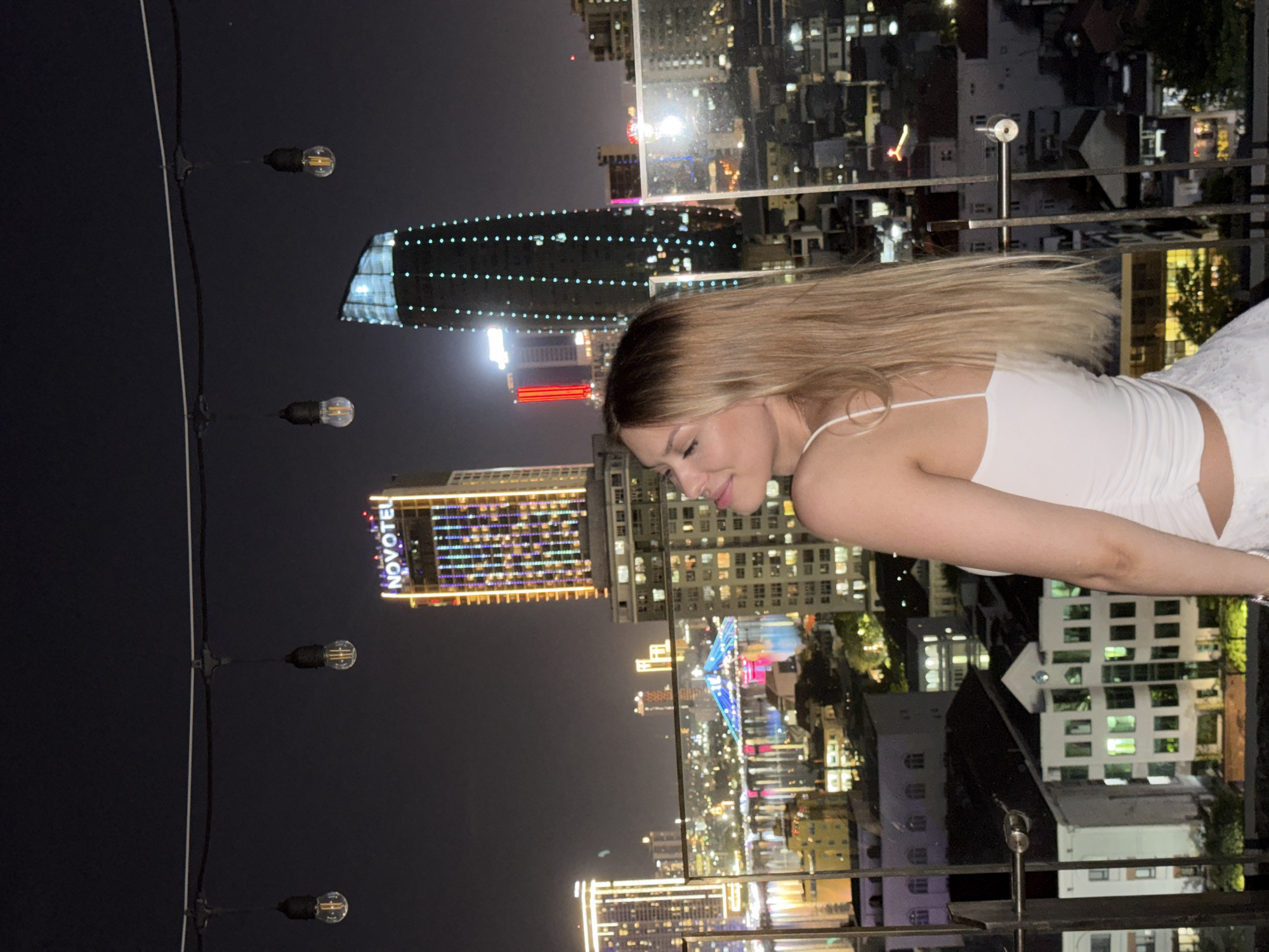 A young woman with blonde hair is captured in a medium shot, standing on a rooftop overlooking the city of Đà Nẵng, Vietnam at night. She is wearing a white tank top and white shorts. The city skyline is illuminated by numerous lights, with tall buildings and a bridge visible in the background. Several string lights hang above the woman, and the scene is dark, suggesting it is nighttime. The woman is looking away from the camera, her head tilted upwards.