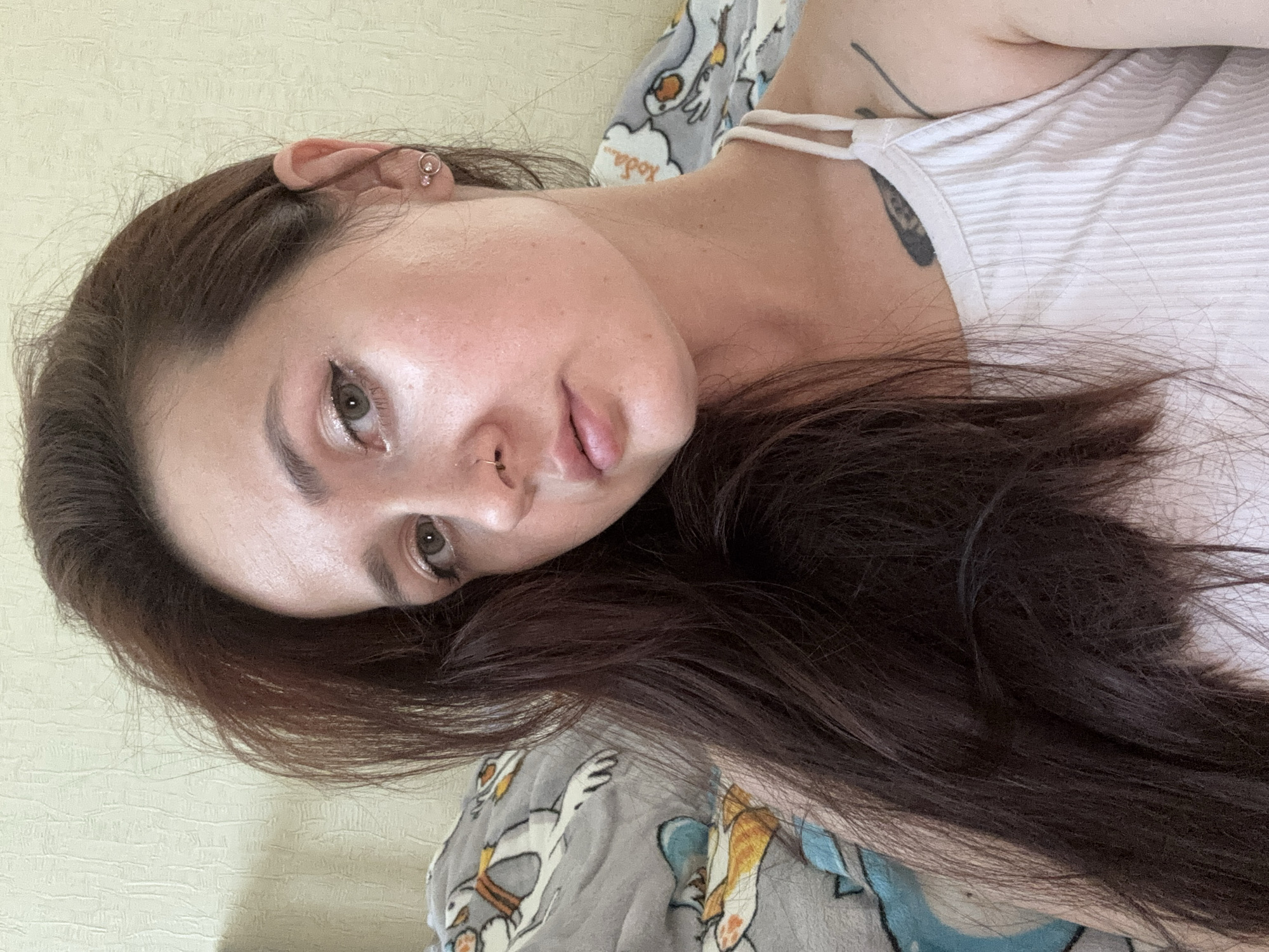 A medium close-up selfie of a young woman with long, dark brown hair and light skin is captured from the chest up. She is looking directly at the camera with a neutral expression. Her hair is parted on the left and falls across her face and chest. She is wearing a light-colored, ribbed tank top. She has a nose ring and small earrings. There is a tattoo on her left shoulder blade. The background consists of a textured, pale yellow wall and a blurred blanket with a cartoonish pattern featuring birds. The scene appears to be indoors, possibly in a bedroom, and the lighting suggests it is daytime. The image does not contain any specific landmarks or elements that would indicate a location within Naberezhnye Chelny, Russia.