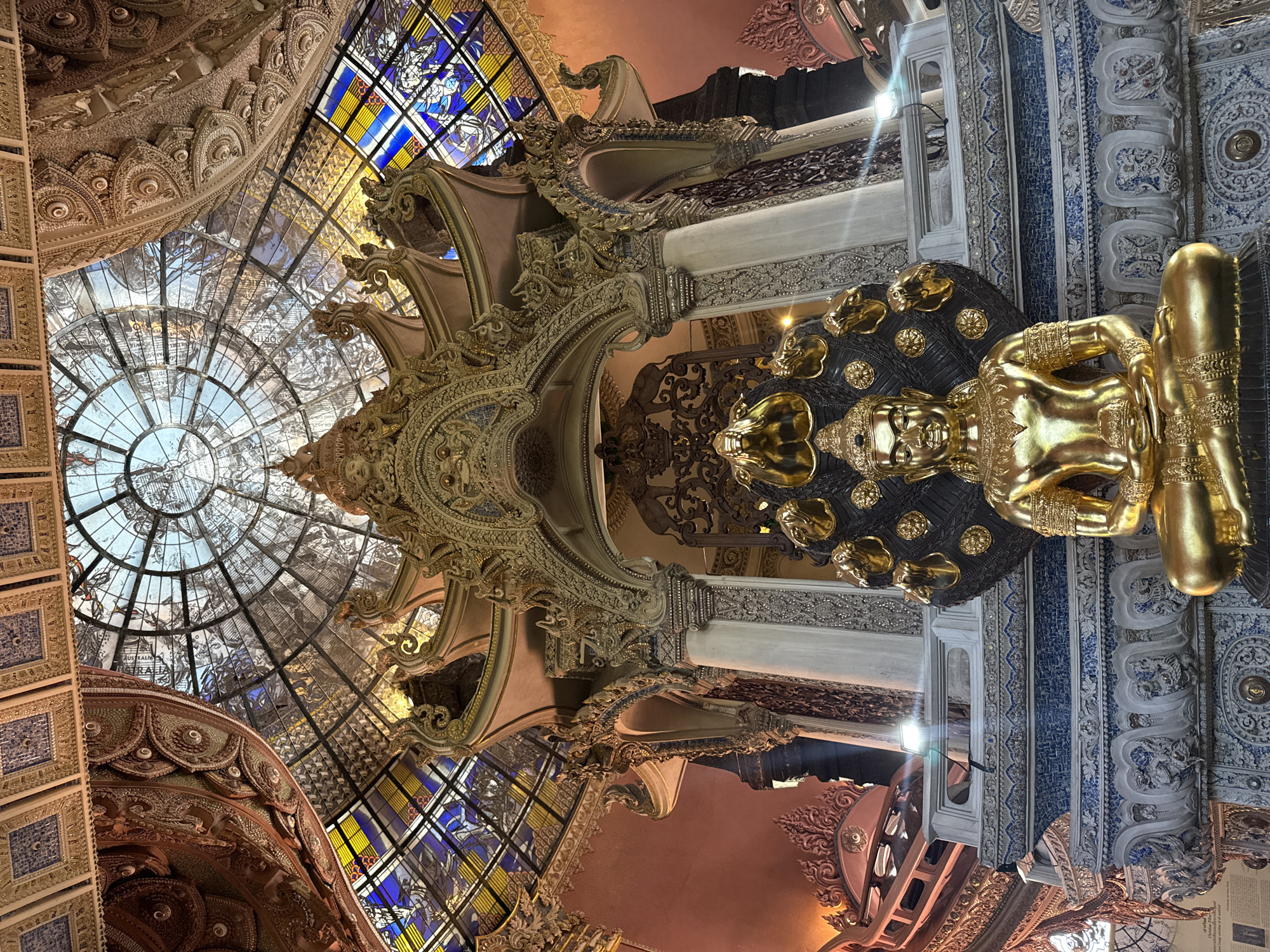 The image depicts an ornate interior space in Samrong Nuea Subdistrict Municipality, Thailand, featuring a golden reclining Buddha statue. Above the statue, there's a decorative ceiling with a glass dome displaying map-like designs and stained-glass windows with blue, yellow, and white elements. The architecture includes intricate carvings and moldings with gold accents. The pillars and beams feature detailed patterns and designs.