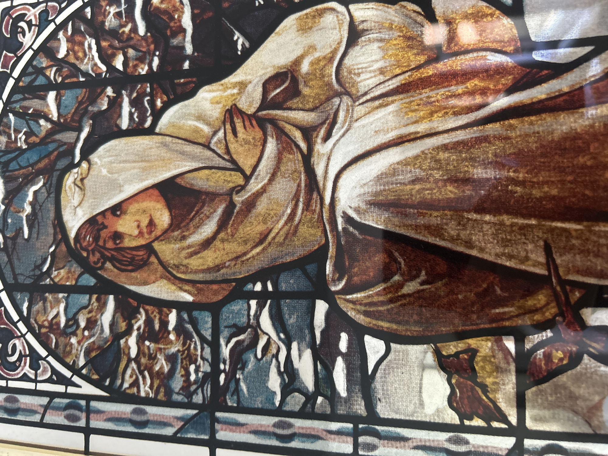The image shows a stained-glass style artwork featuring a woman with her hands clasped in prayer, cloaked in a light-colored robe with gold highlights. She is surrounded by an abstract nature scene, consisting of trees and branches in blues, browns, and whites. The artwork is framed by a decorative glass border featuring stylized floral and geometric motifs. The exact location of the artwork in Câmara de Lobos, Portugal is not discernible from the image alone.