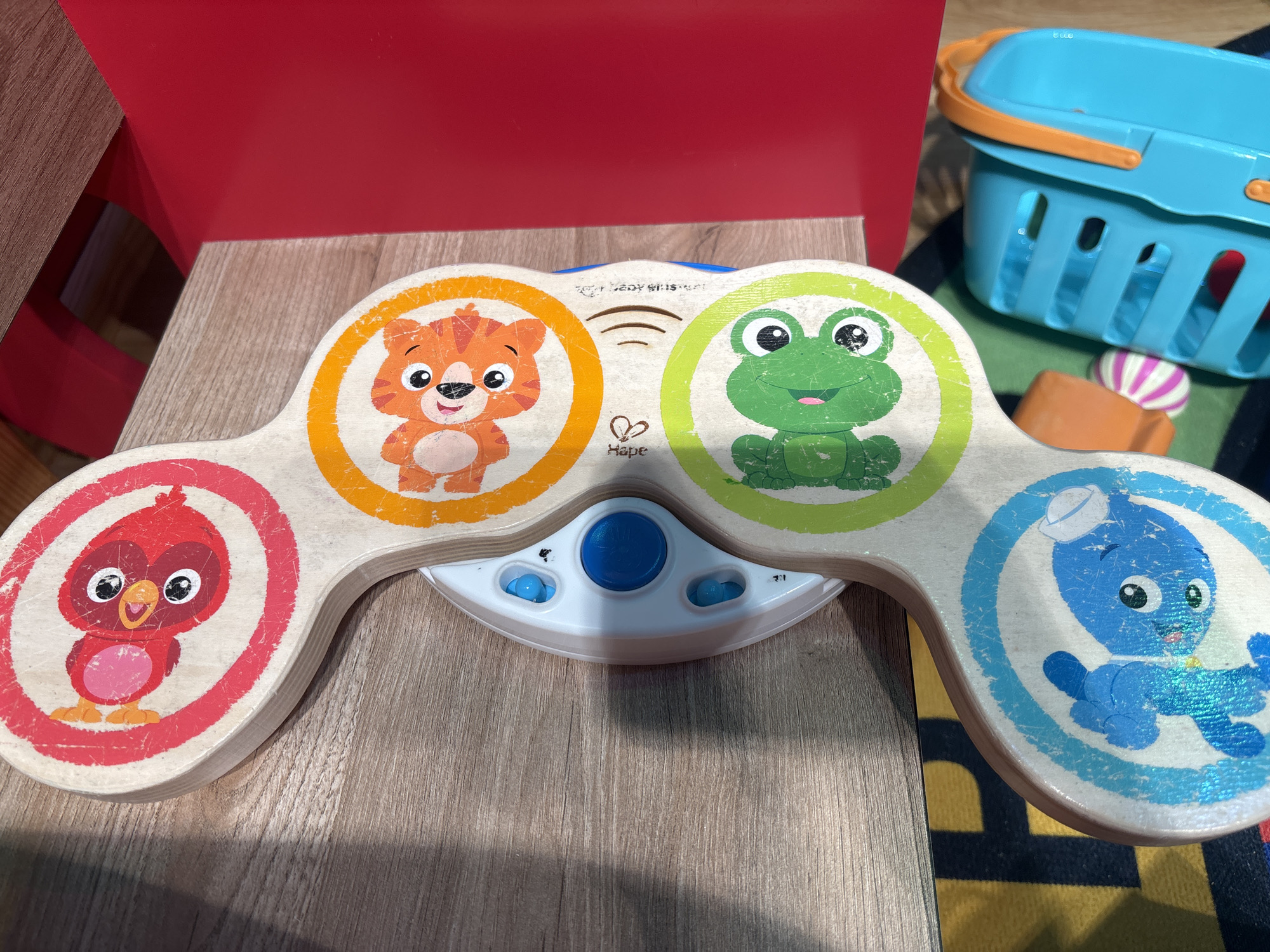A wooden toy drum set with images of animals is placed on a wooden table, with a red chair in the background. The toy has four circular sections, each depicting a different animal: a red bird at the top, an orange tiger next, a green frog below that, and a blue dog at the bottom. In the center of the toy are white plastic components with blue buttons, likely for sound or light functions. The toy appears to be made by the brand Hape, as indicated by a logo. To the right of the toy, a bright blue plastic toy basket with orange handles is visible, along with a small, striped ball. The floor beneath the table has a colorful rug with black and yellow stripes. The scene is indoors and appears to be in a home or play area. There are no people present in the image. The lighting suggests artificial indoor lighting. The image was taken in Dubai, United Arab Emirates.
