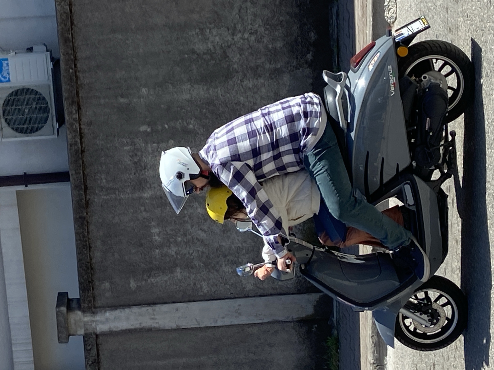Two individuals are riding a gray Veracruz scooter on what appears to be a paved street. The driver, in front, wears a white helmet, a purple and white checkered shirt, and blue jeans. The passenger, behind, wears a yellow helmet and a white jacket. The scooter is a gray Veracruz model. The location includes a building with a Midea air conditioner unit affixed to its wall. The specific location is Budva, Montenegro, due to scooter brand/availability. The scooter appears to be stationary or moving very slowly.