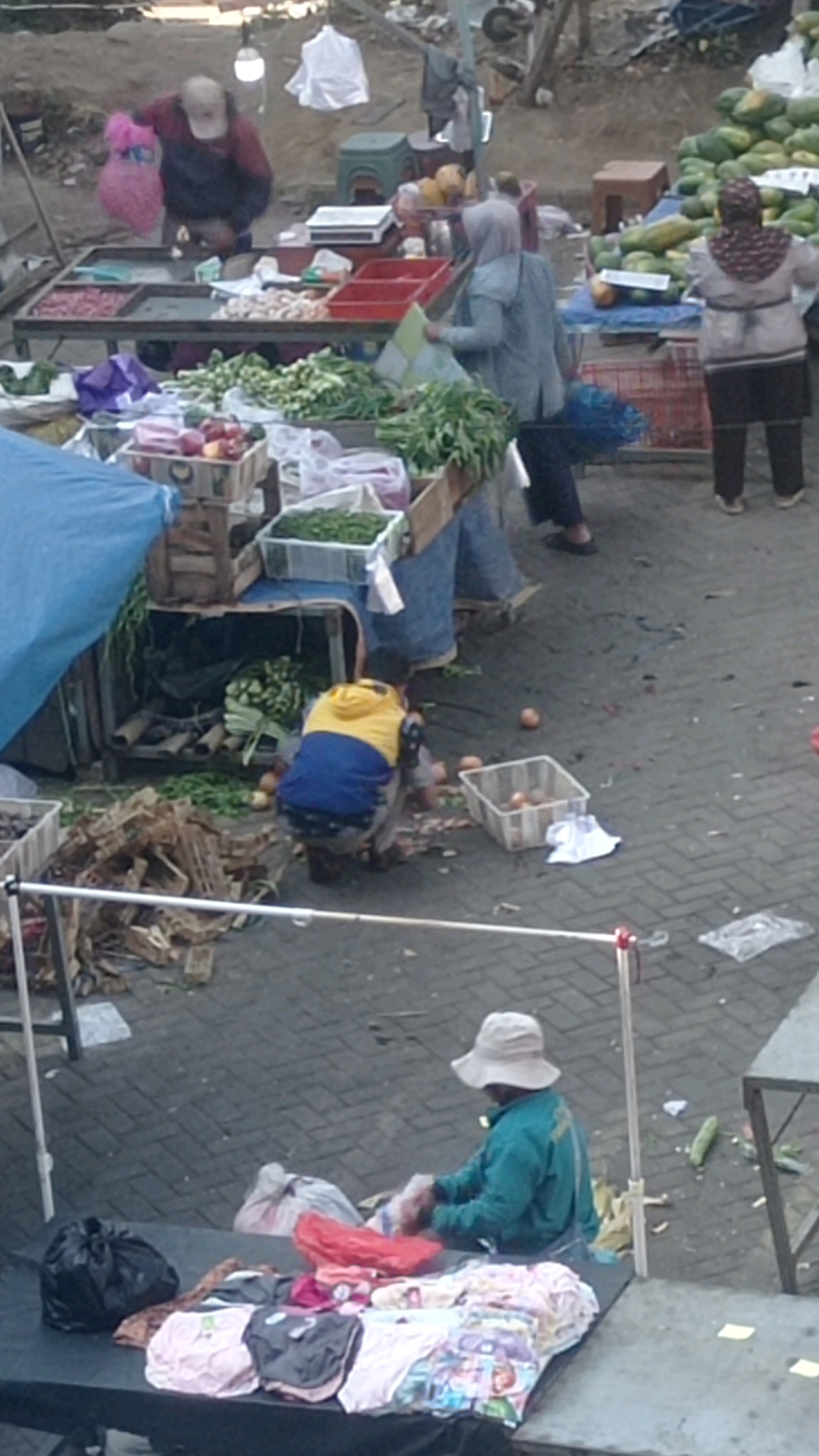 This is an eye-level, medium shot, taken outdoors at a street market in Batu, Indonesia. The scene depicts an outdoor market with several individuals engaged in various activities. In the foreground, a person wearing a wide-brimmed hat and a turquoise jacket is sitting behind a table laden with folded clothing, appearing to be arranging or sorting items. A black plastic bag and a red plastic bag are also on the table. Behind this individual, to the left, is a pile of what appears to be discarded wooden material.
Further back in the scene, another person in a blue and yellow jacket is crouched down, interacting with produce on the ground. Several individual onions are scattered on the paved ground around them, along with a plastic crate containing more onions and some scattered debris. In the mid-ground, there are market stalls with various produce, including leafy greens, papayas, and what appear to be onions or garlic, displayed in crates and on tables. Several people, some wearing headscarves, are visible near these stalls, with one person holding a pink bag. The ground is paved with interlocking bricks. The overall environment suggests a busy, open-air market.