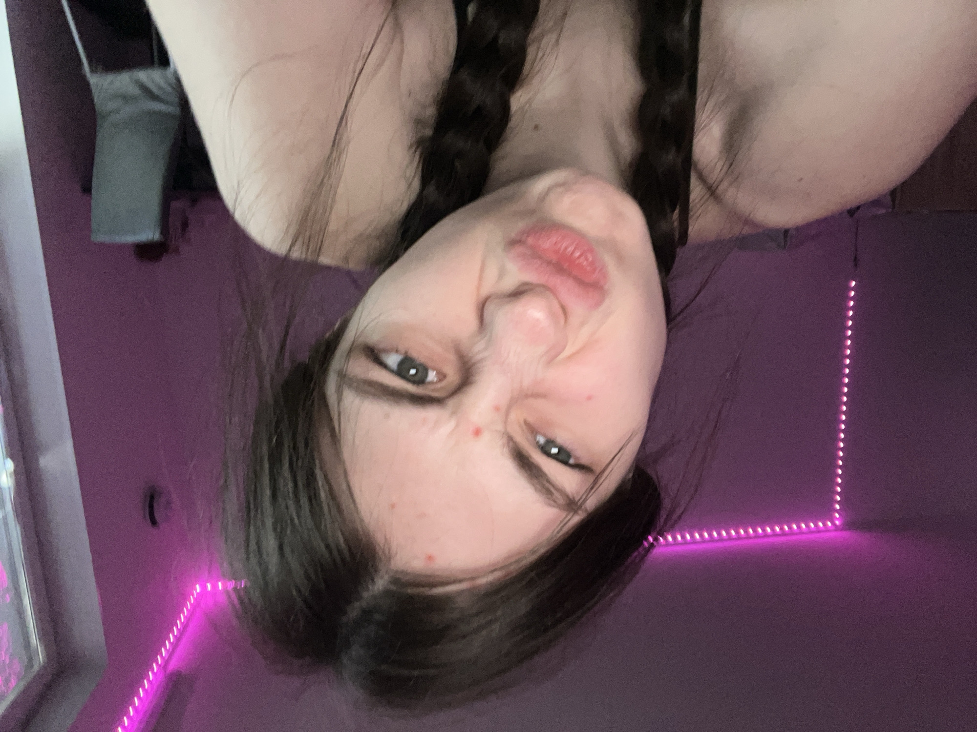 A young woman with light skin, dark brown hair braided into two pigtails, is facing the camera with her face scrunched up. She has green eyes and her lips are pursed. The background is a purple wall with pink LED strip lights running along the corner of the room. A gray fabric object hangs in the upper left corner. The image is rotated upside down. The location and city are not discernible.