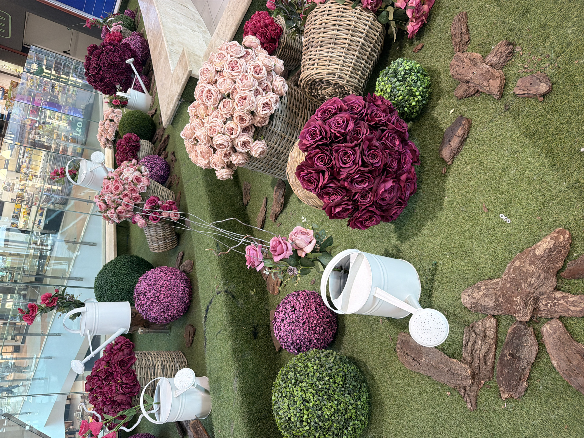 This indoor scene in Sofia, Bulgaria shows a floral display on a grassy surface. Several bouquets of pink and burgundy roses are arranged in woven baskets, interspersed with green and purple spherical bushes. White watering cans are scattered throughout. Wood bark pieces are also present on the grass. The background reflects a multi-level store with glass facades.