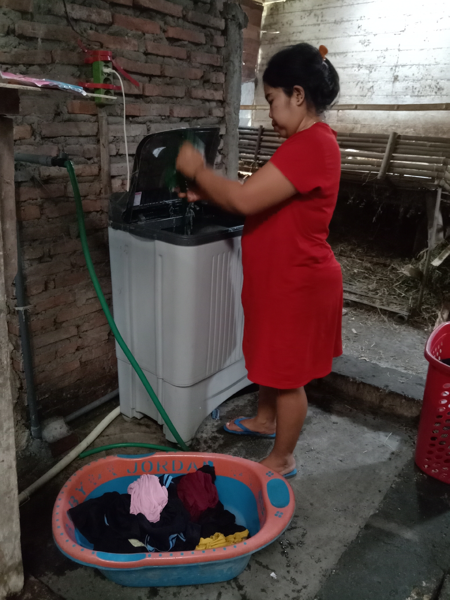 In the city of Jumput, Indonesia, a woman in a red dress is standing next to a white washing machine and a large plastic basin filled with laundry. She appears to be in the process of washing clothes. The washing machine is connected to a green hose, suggesting it's being filled with water. The background shows a brick wall, wooden structures, and dry vegetation, indicating an outdoor or semi-outdoor setting, possibly a laundry area. A red laundry basket is visible to the right of the scene.