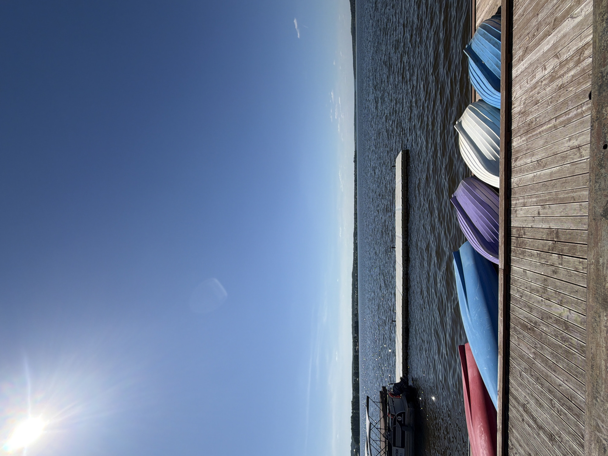 The image depicts a wooden dock extending into a body of water under a clear blue sky. The sun is visible in the lower left corner, causing a glare. Docked alongside the platform are several overturned kayaks in varying colors including blue, white, purple, blue again, and red. The body of water stretches to the horizon and it appears to be Lake Votkinsk in Votkinsk, Russia. There is a floating platform extending out into the lake with a small boat docked alongside.