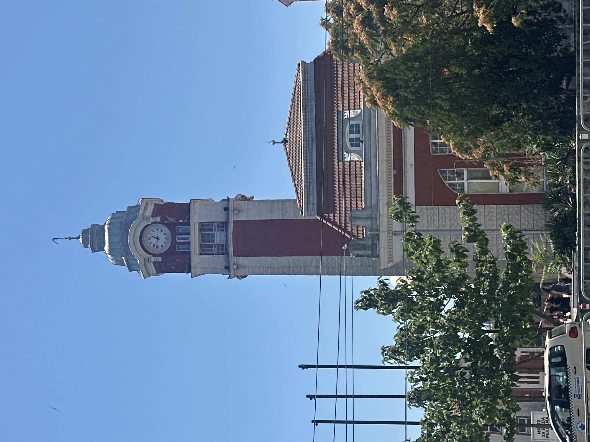 This is a vertical, medium shot of a clock tower and building under a clear blue sky. The clock tower is tall and has a red and white facade with a grey, domed top. The clock face is visible and shows the time as approximately 10:37. Below the clock tower is a red and white building with a red tiled roof. In the foreground, there are trees and what appears to be a street with a white car and some people walking. Overhead power lines are also visible.