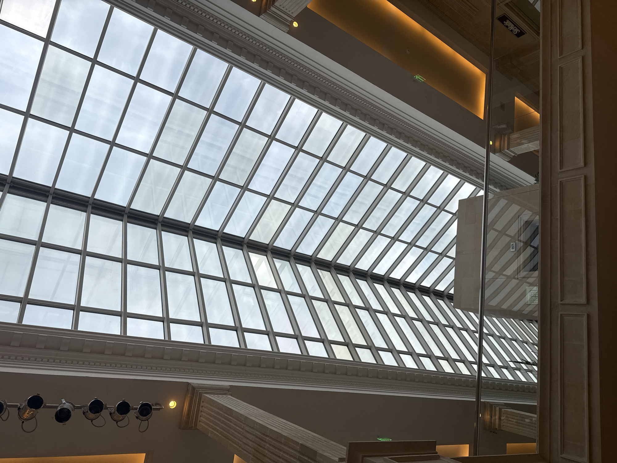The image shows an interior view looking up towards a large, angled glass ceiling. The ceiling is divided into a grid of rectangular panes by a dark framework. Through the glass, a cloudy, overcast sky is visible, suggesting it is daytime but with diffused light.
Below the glass ceiling, there is ornate architectural detailing, including moldings and columns. In the lower part of the image, a row of spotlights is mounted on a surface, pointing upwards or outwards. To the right, a tall, reflective glass pillar stands vertically, showing a distorted reflection of the ceiling and interior space.
The overall setting appears to be a grand indoor space, possibly a hall, museum, or atrium, characterized by its high ceiling and decorative elements. There is no activity or event depicted, and the scene is static. No people are visible. The lighting from above the ceiling is warm and diffused. There is a small green sign with white text visible on the ceiling. The text on the reflective pillar appears to be a reflection of a sign that reads "Selfridge's". The time of day is likely daytime due to the natural light, but the weather is overcast. The emotions conveyed are neutral and architectural.